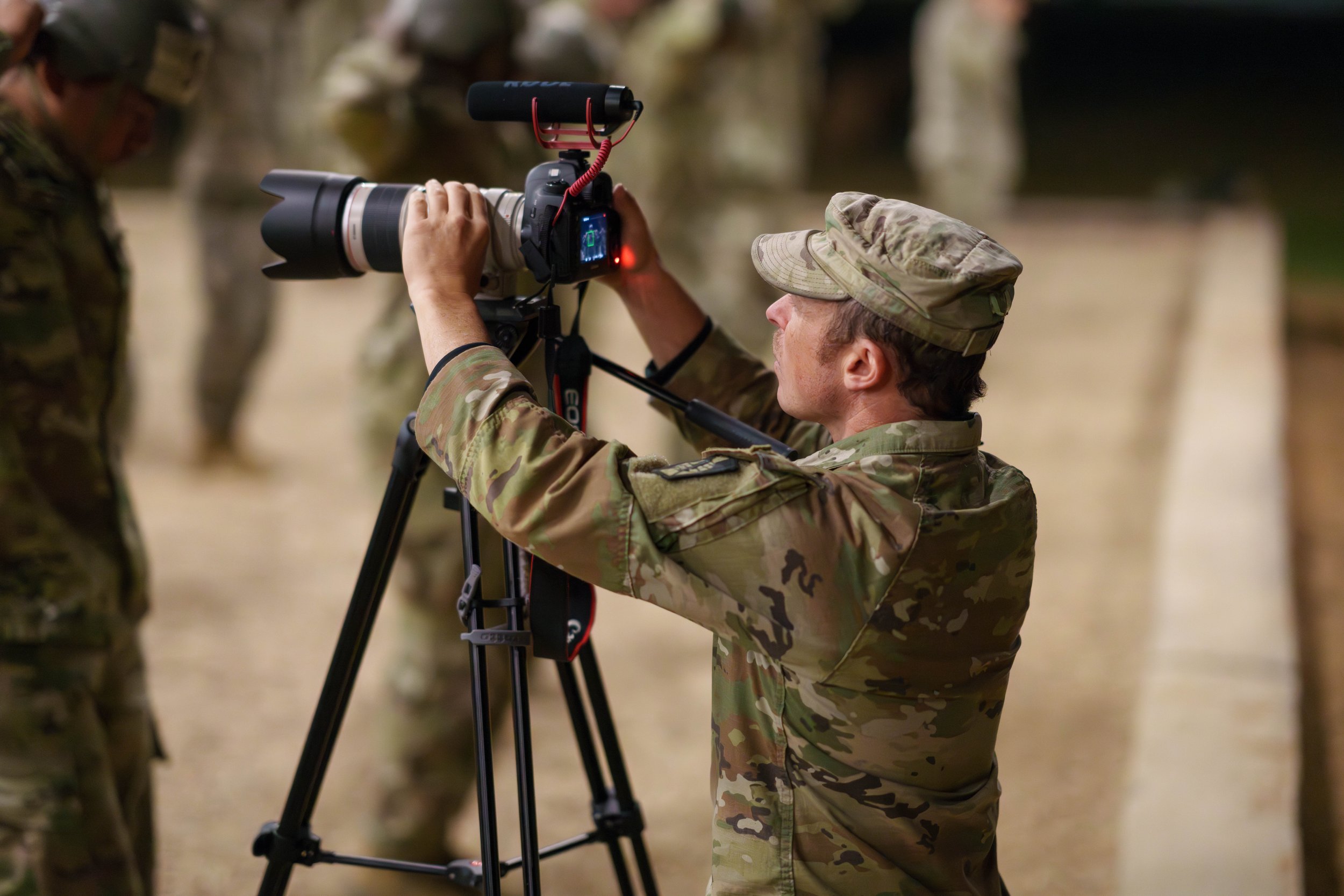 A soldier in camouflage uniform adjusting a camera on a tripod, with other soldiers in the background.