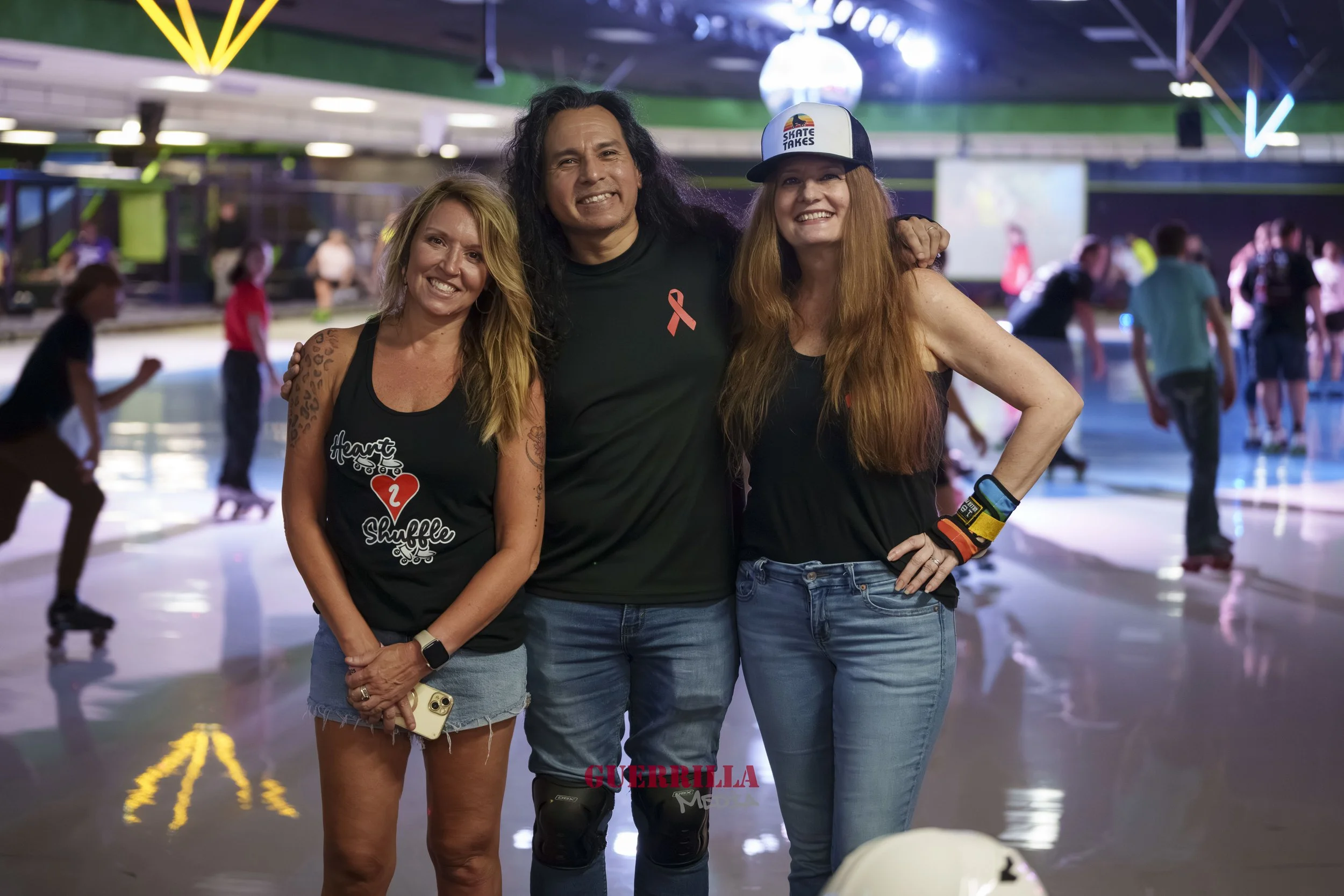 Three smiling women and a man at a roller skating rink, with other skaters in the background. They are standing together for a photo, some wearing wrist guards and casual clothing.