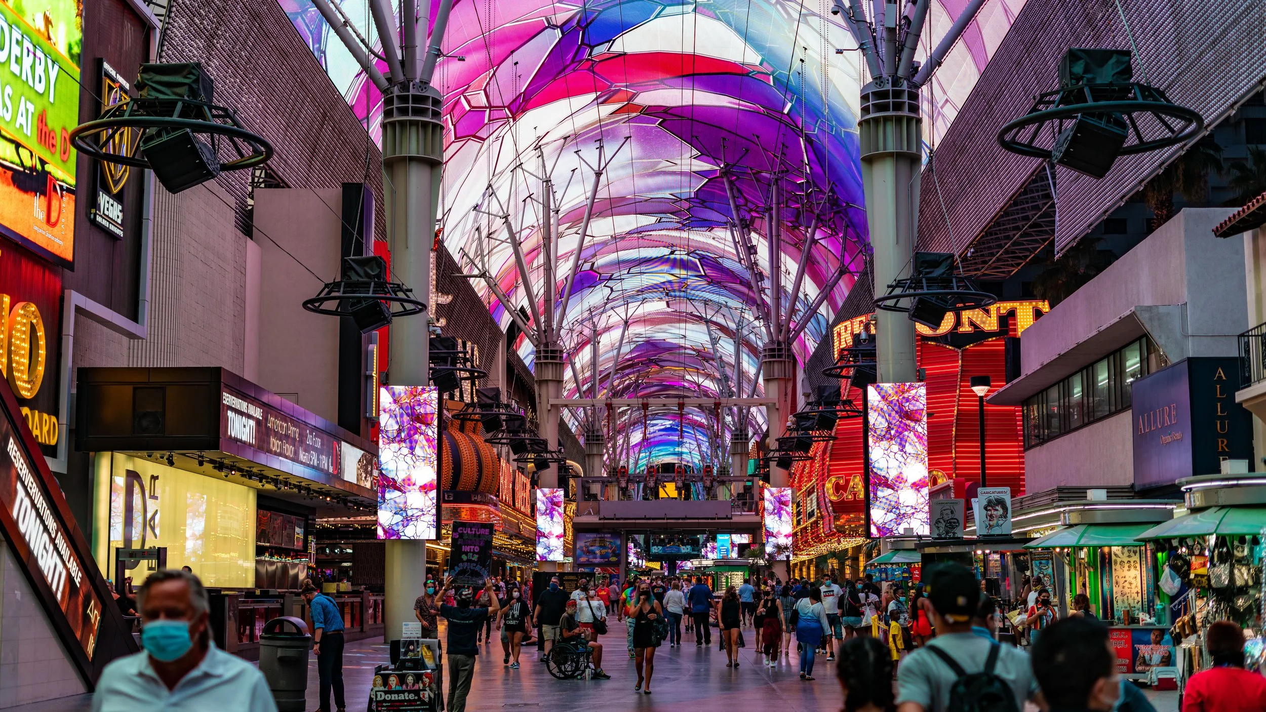 Crowded shopping area under a colorful, illuminated canopy with bright signs, LED screens, and street vendors.
