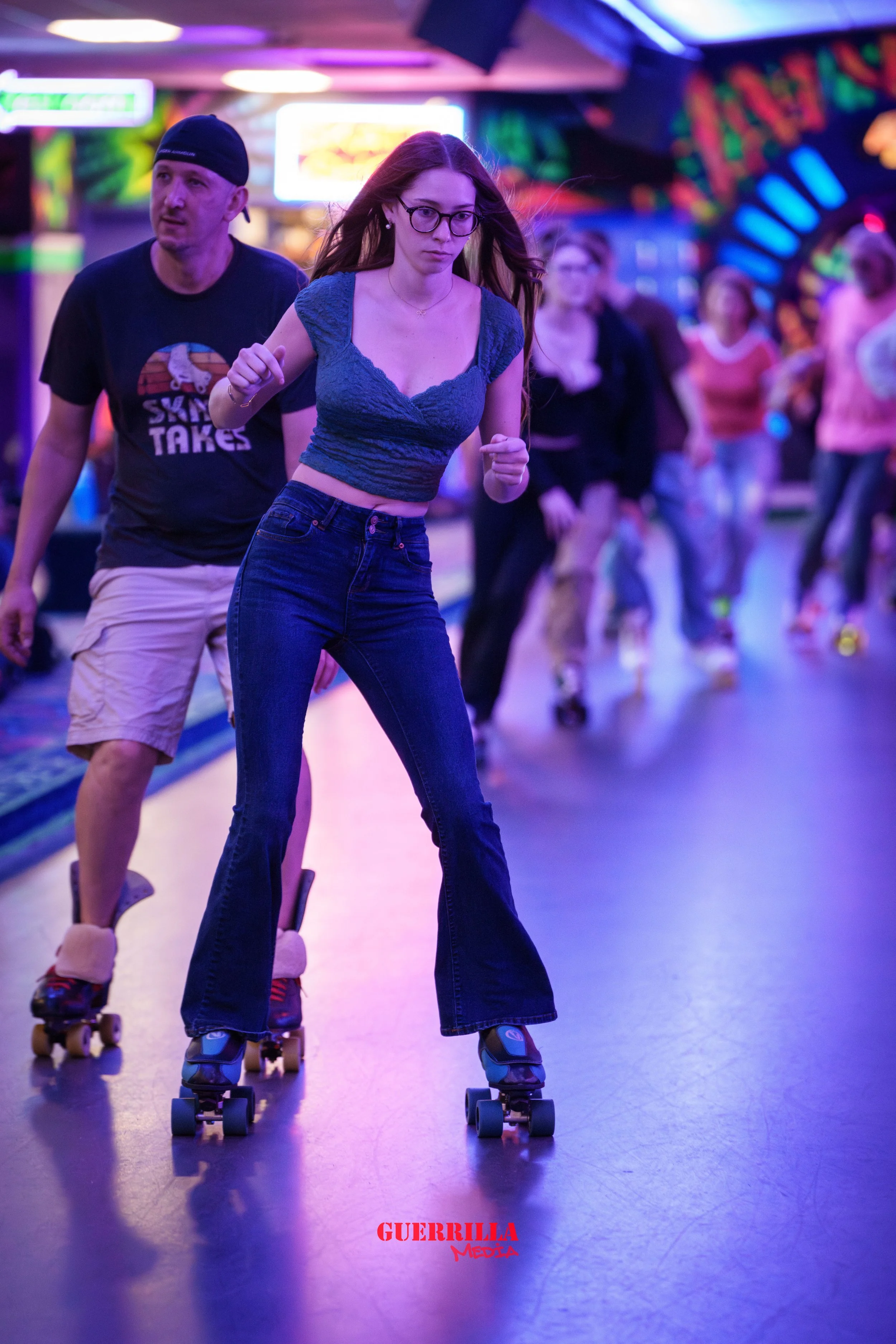 Young woman roller skating on a roller rink, wearing glasses, a blue crop top, and high-waisted jeans, with a man roller skating behind her, surrounded by other skaters in the background, lit by colorful neon lights.