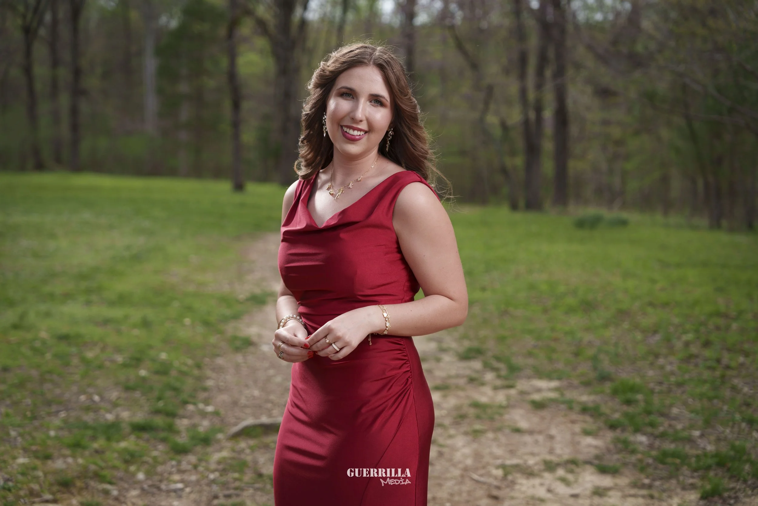 A woman in a red dress standing on a dirt path in a wooded park area with green grass and trees, smiling at the camera.
