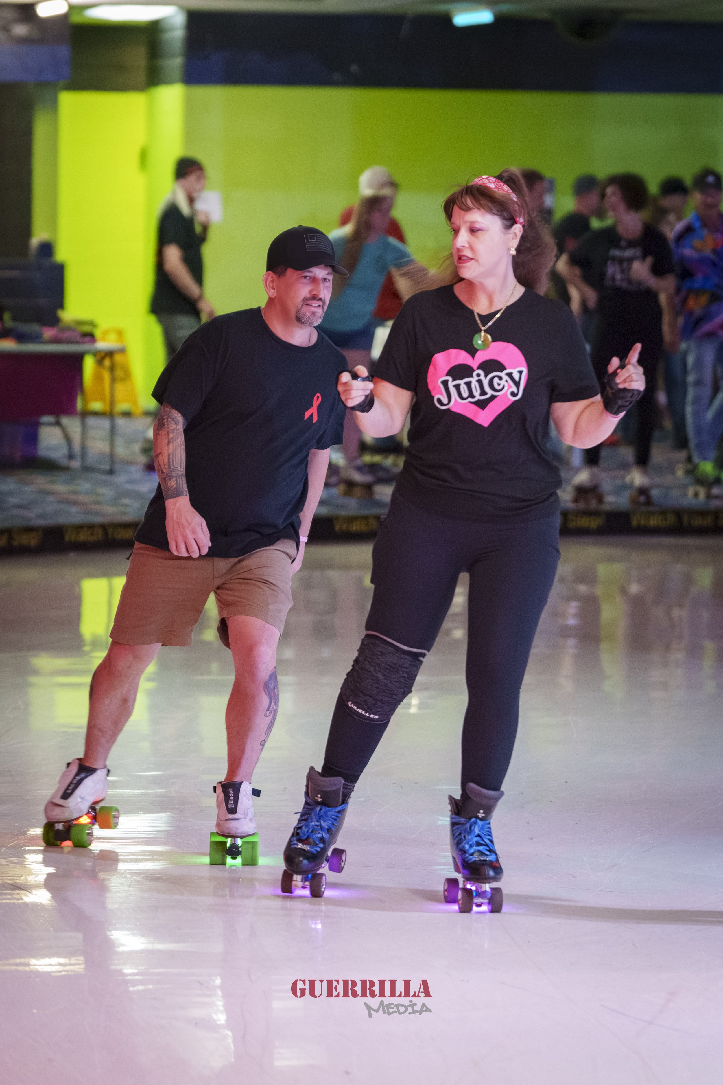 Two people roller skating indoors, one man and one woman, with a colorful background and other skaters in the distance.