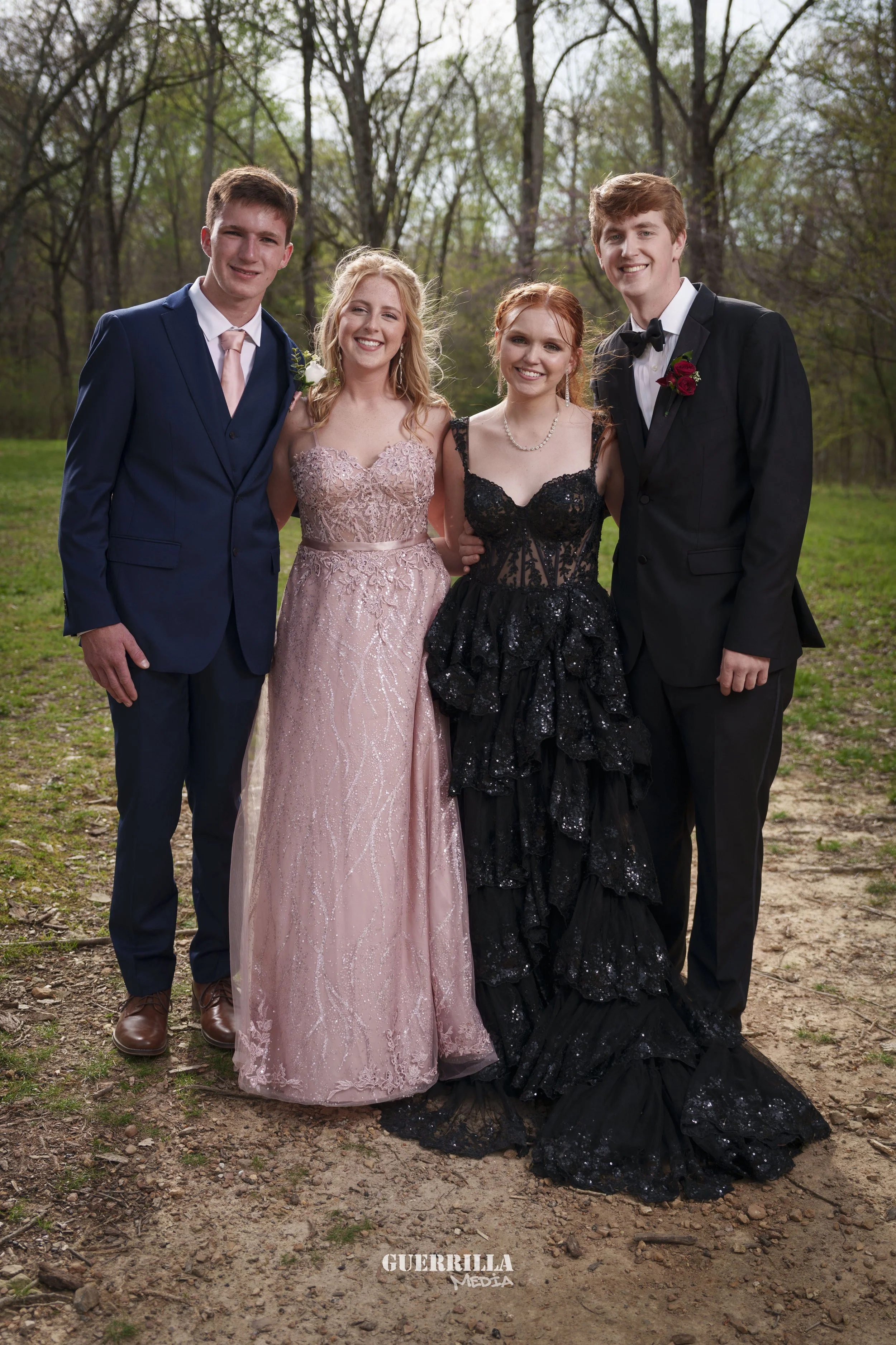 Four young adults dressed in formal attire standing outdoors in a wooded area, smiling for a photo.
