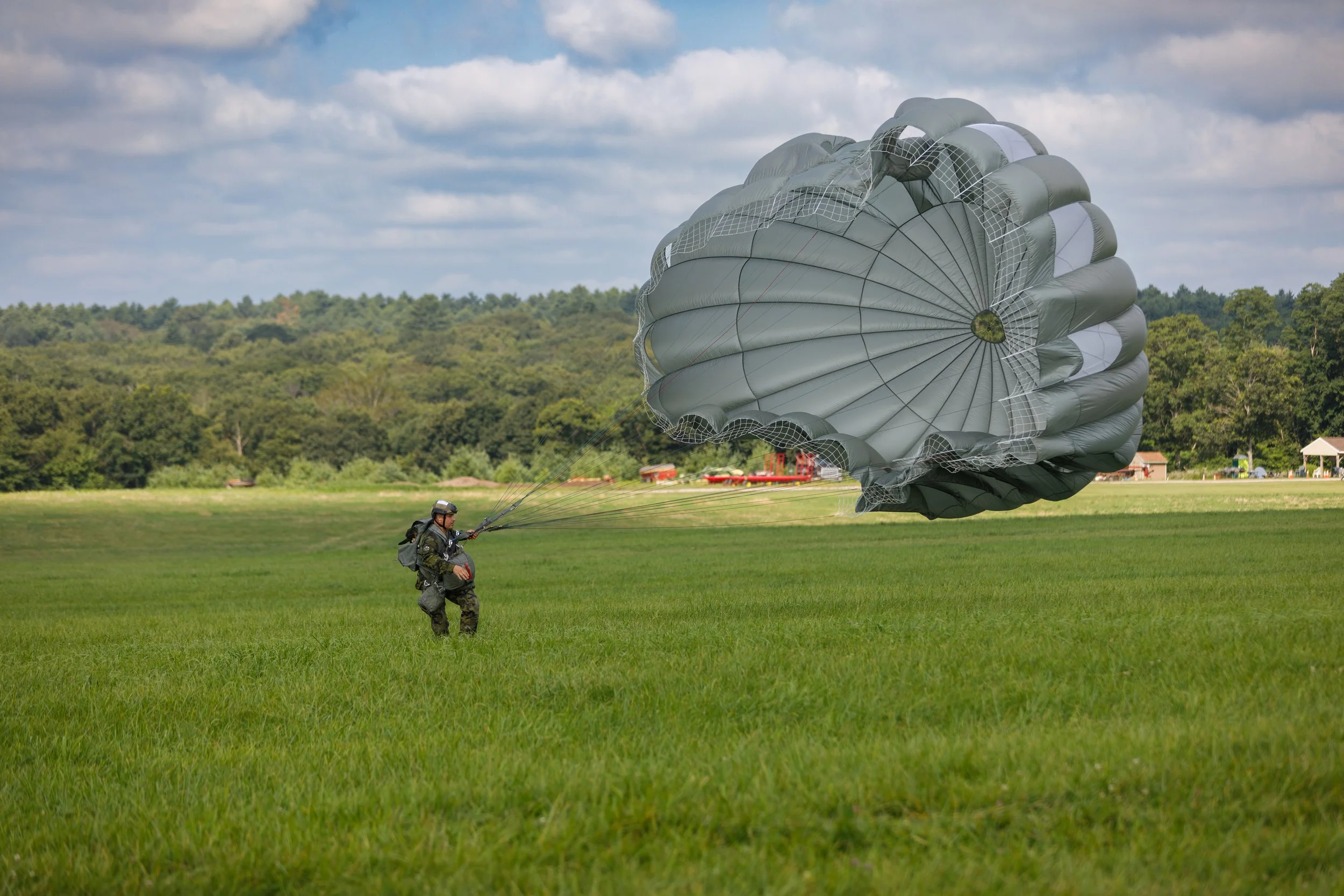 A soldier preparing to land with a parachute on a grassy field, with trees and a cloudy sky in the background.