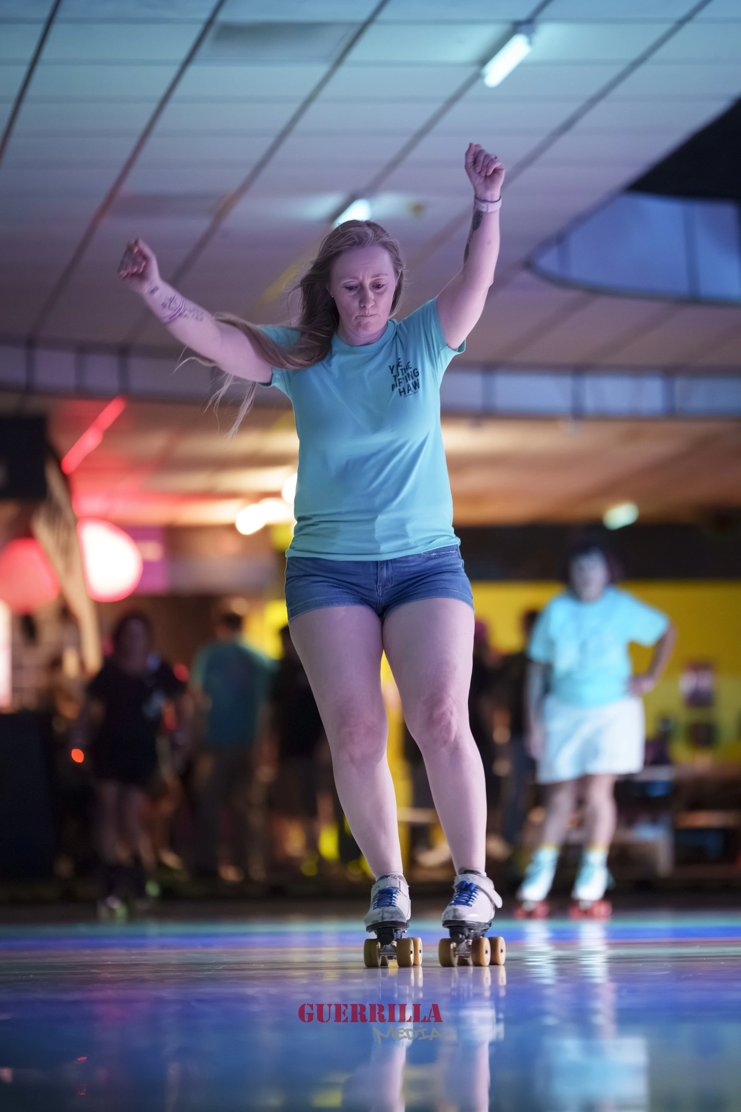 A woman in a blue shirt and denim shorts roller skating indoors, with her arms raised and a focused expression. There are others in the background and colorful lights in a roller rink setting.