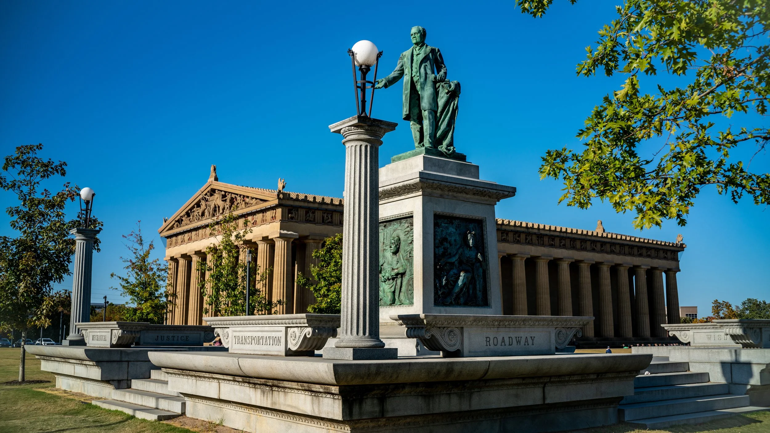 Statue of a man standing on a pedestal with various carvings, in front of a classical building with columns, trees, and clear blue sky.