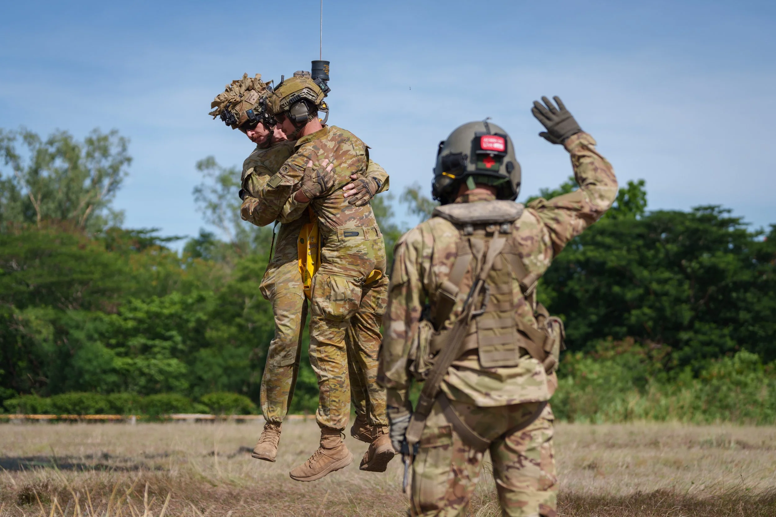 Two soldiers are airborne, embracing each other mid-air, while a third soldier on the ground watches and raises his hand. All are wearing camouflage military uniforms and helmets.