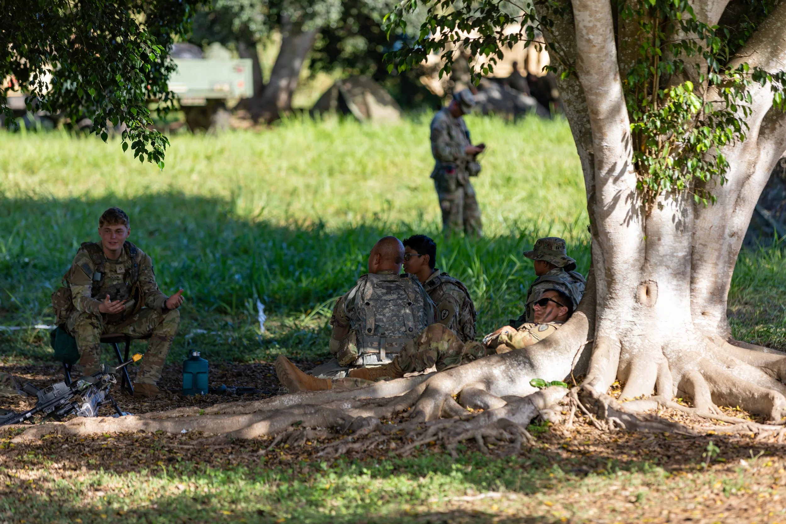 Group of soldiers resting under a large tree with exposed roots in a grassy outdoor setting, some sitting and others standing, engaging in conversation and using electronic devices.
