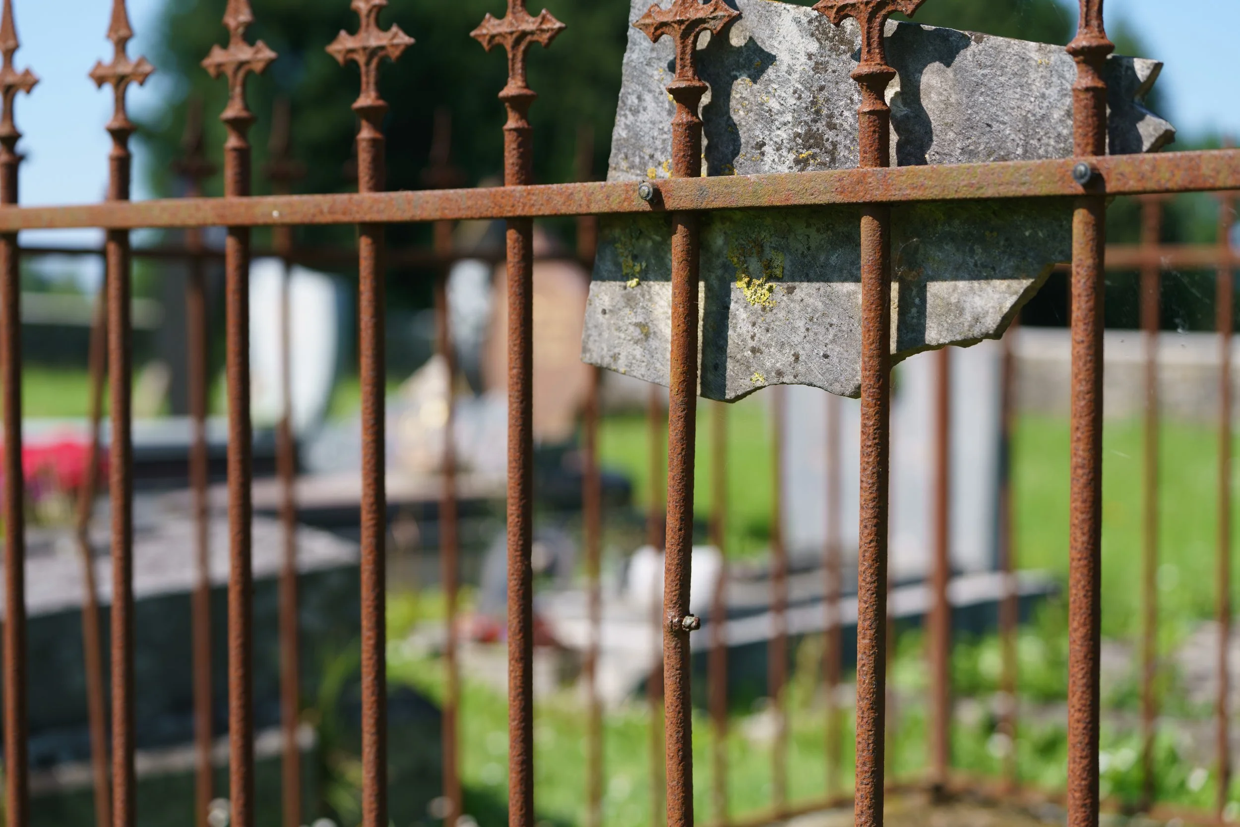 Close-up of a rusty metal fence with a stone behind it, showing moss and lichen, with a blurred background of what appears to be a cemetery with tombstones and green grass.