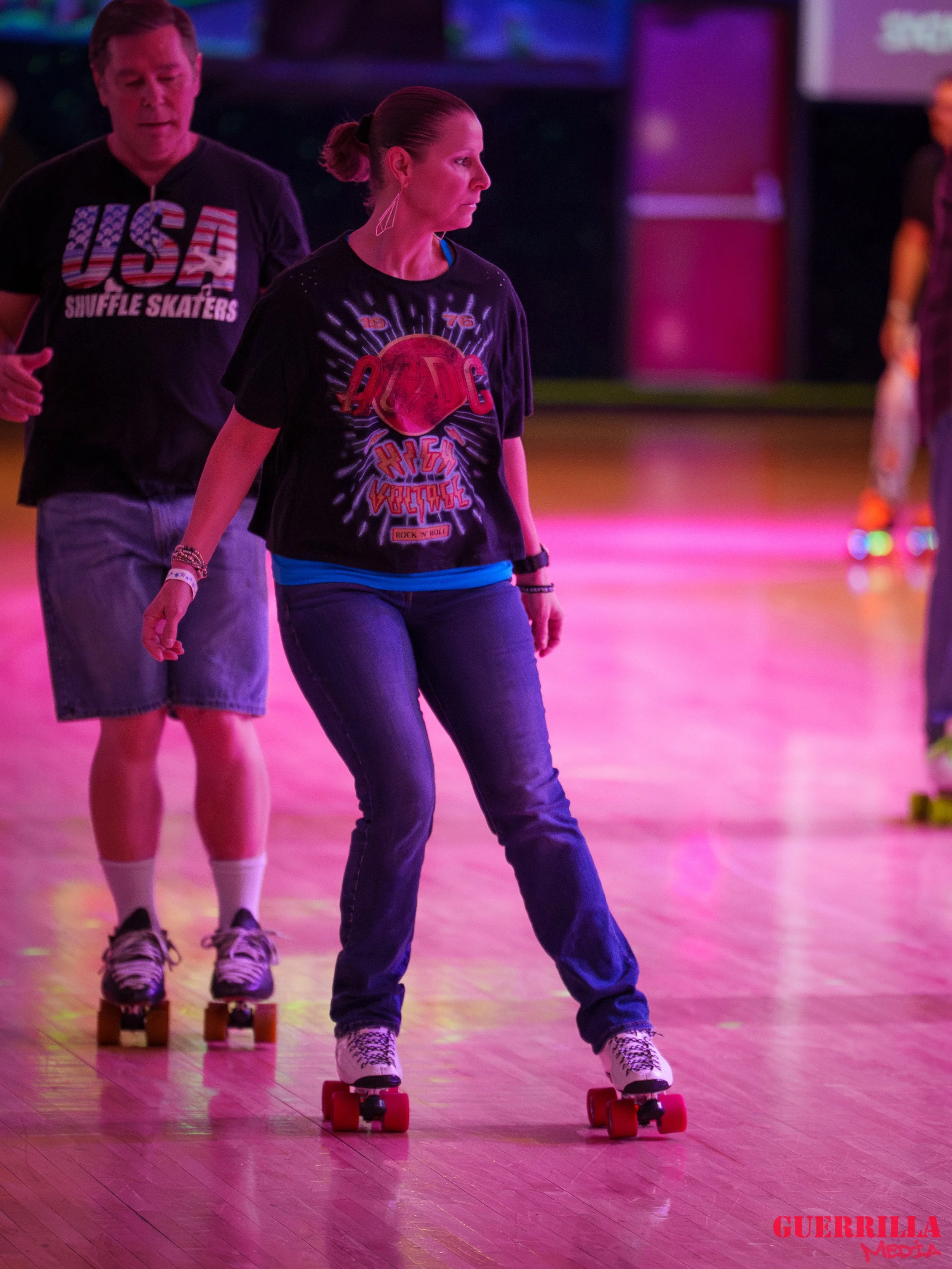 People roller skating indoors under colorful neon lights, with a woman in the foreground wearing a black graphic t-shirt and jeans, and a man behind her wearing a black t-shirt with 'USA Shuffle Skaters' printed on it.