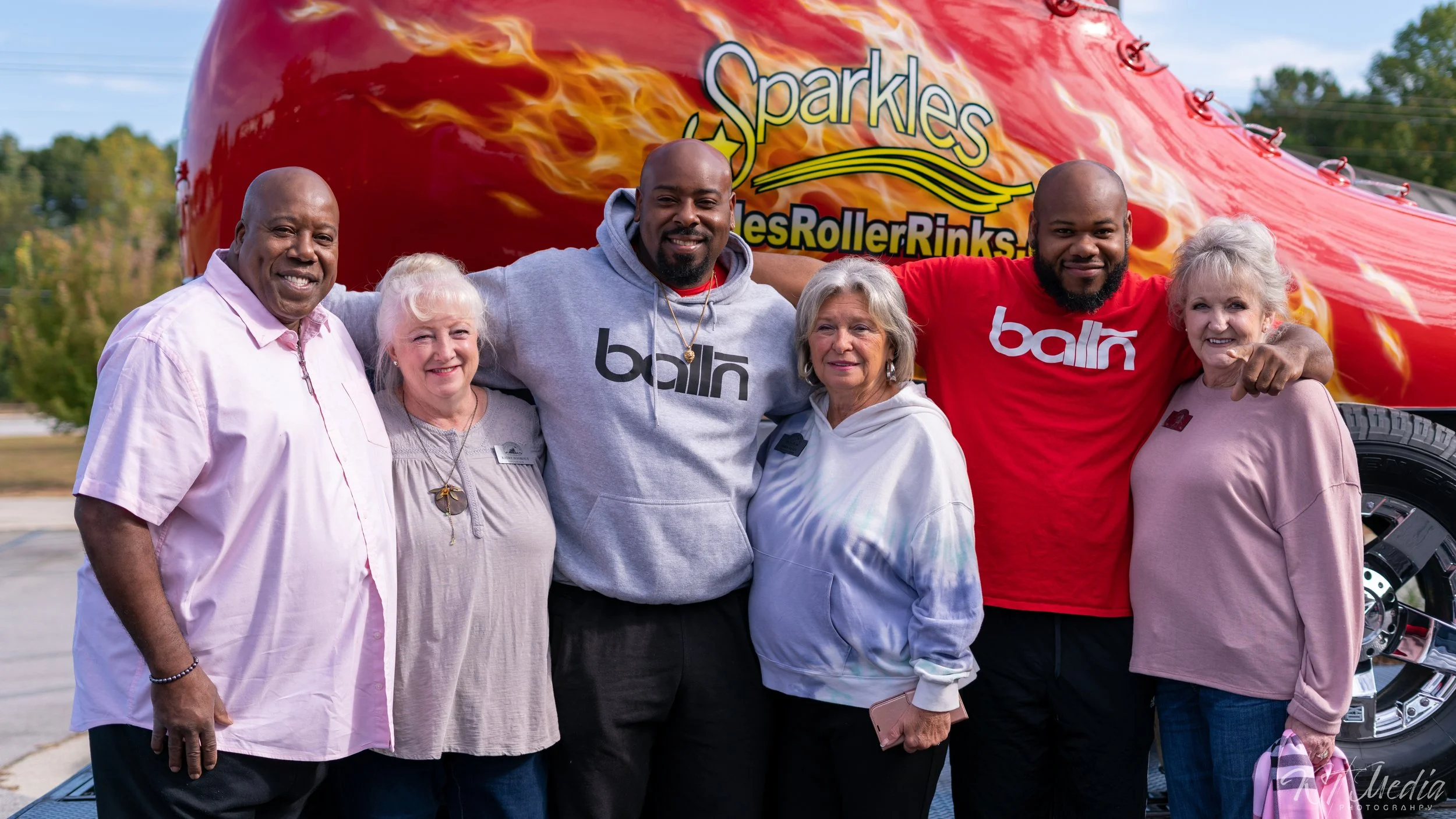 Group of seven people standing together outdoors in front of a red amusement ride with flames and the words "Sparkles" and "Roller Rinks." The group includes four women and three men, some with gray hair, all smiling and embracing each other.