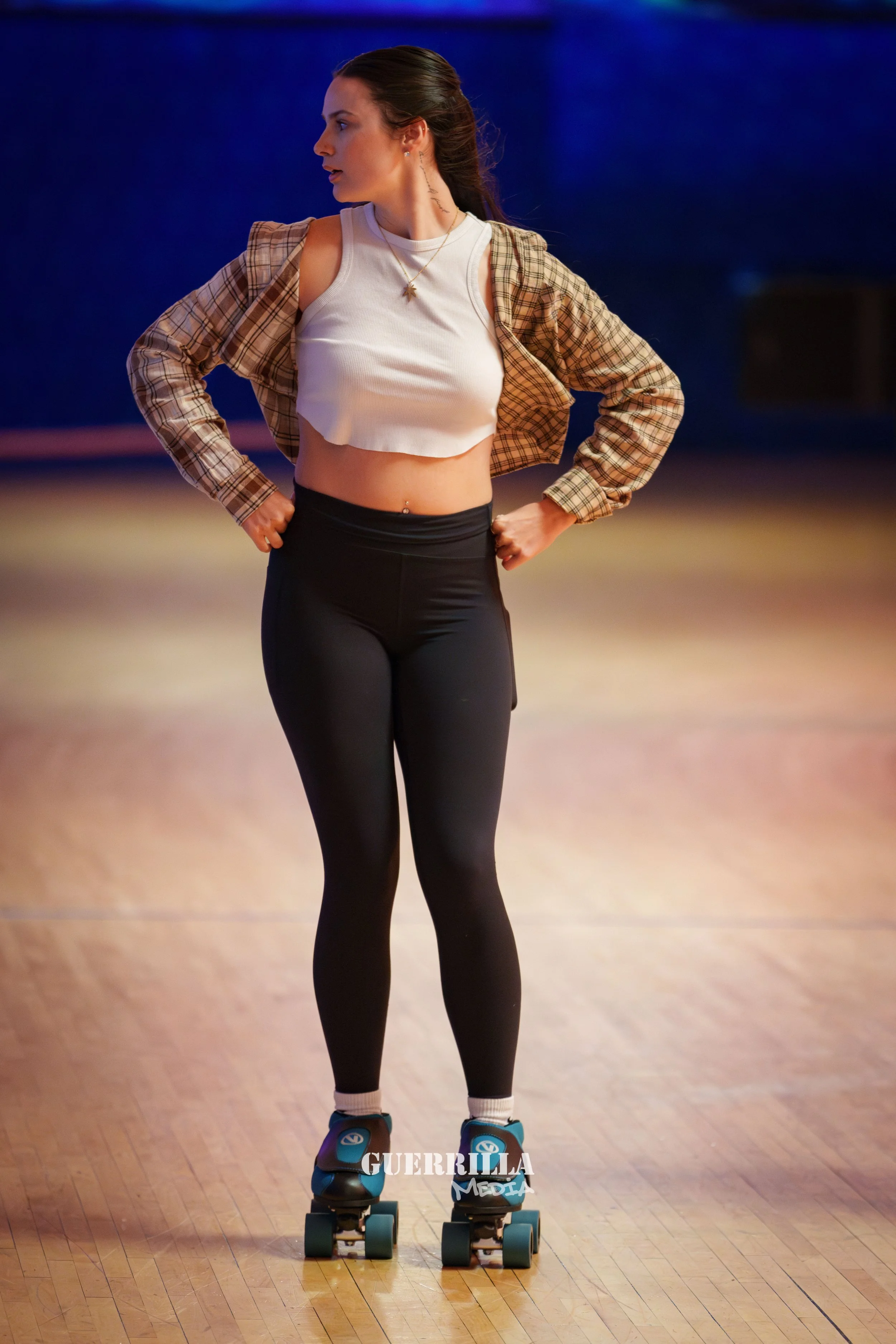 A young woman skating indoors on a wooden floor, wearing a white crop top, black leggings, a plaid shirt, and black skate shoes.