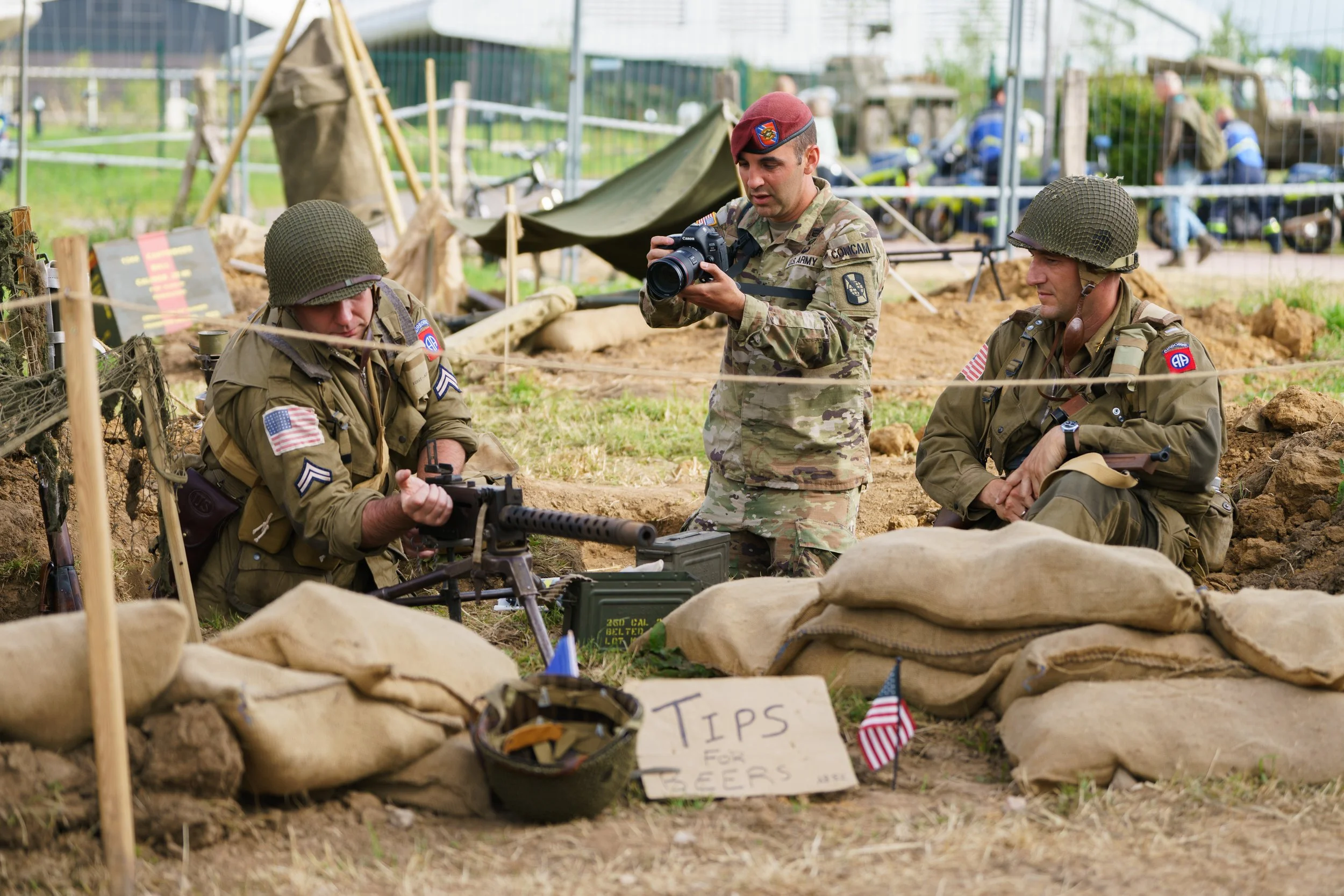 Historical reenactment of soldiers in World War II gear with weapons, sandbags, and American flags, with a sign reading 'Tips for Beers' in front.