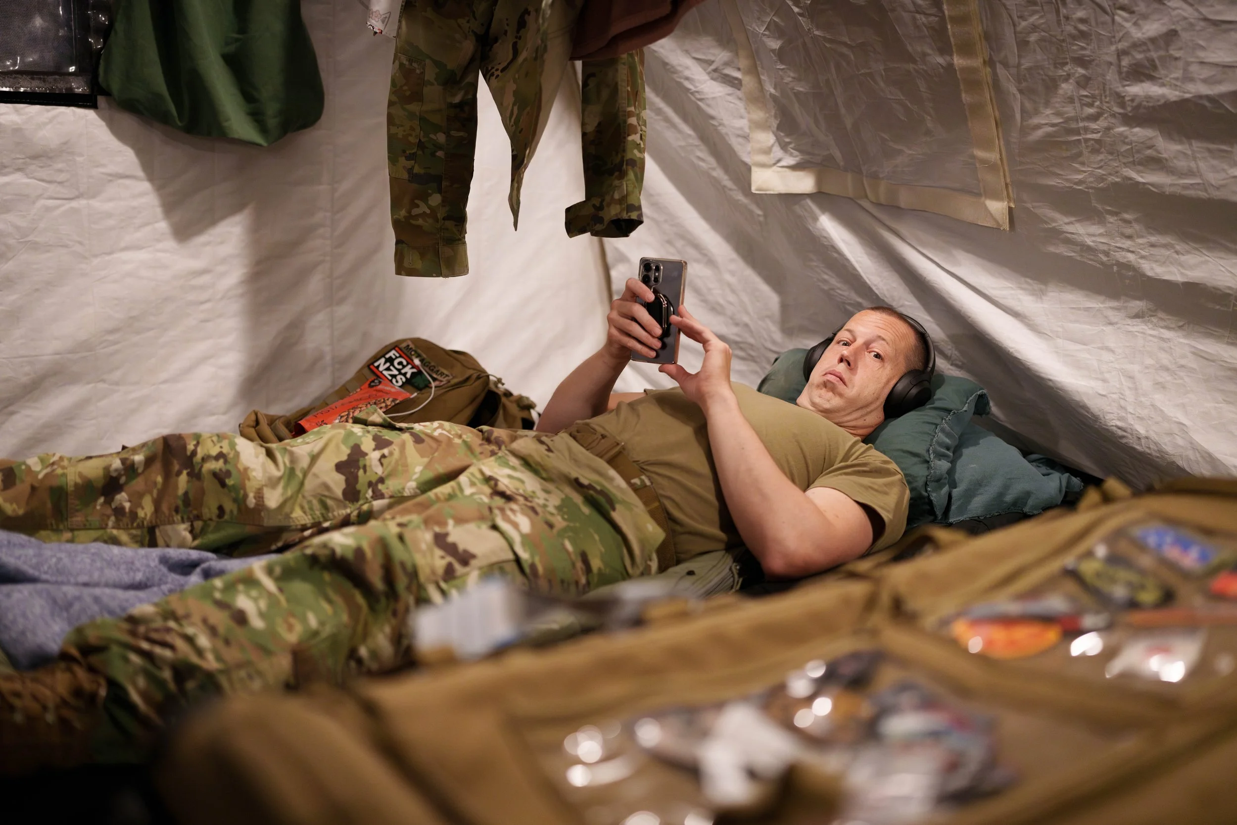 A man in military camouflage uniform lying in a tent, using a smartphone, with camouflage clothing hanging above him and snacks and supplies nearby.