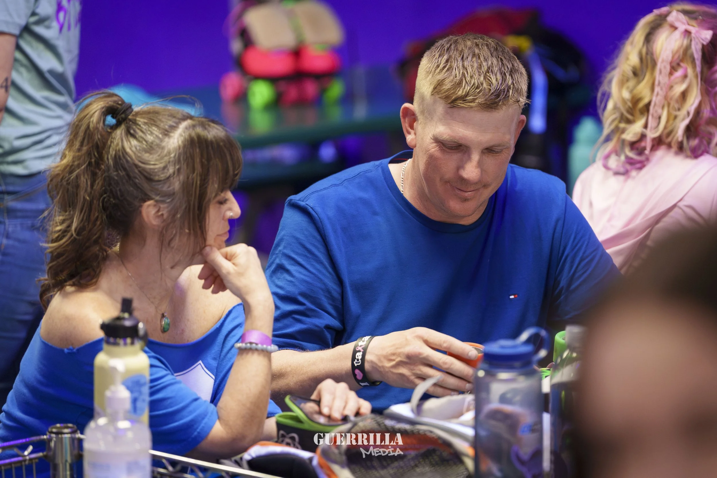 A man and woman sitting at a table in a roller skating rink. The woman is wearing a blue top, has brown hair tied back, and is looking down at the table. The man is also in a blue shirt, blonde hair, and appears to be looking at something in his hand