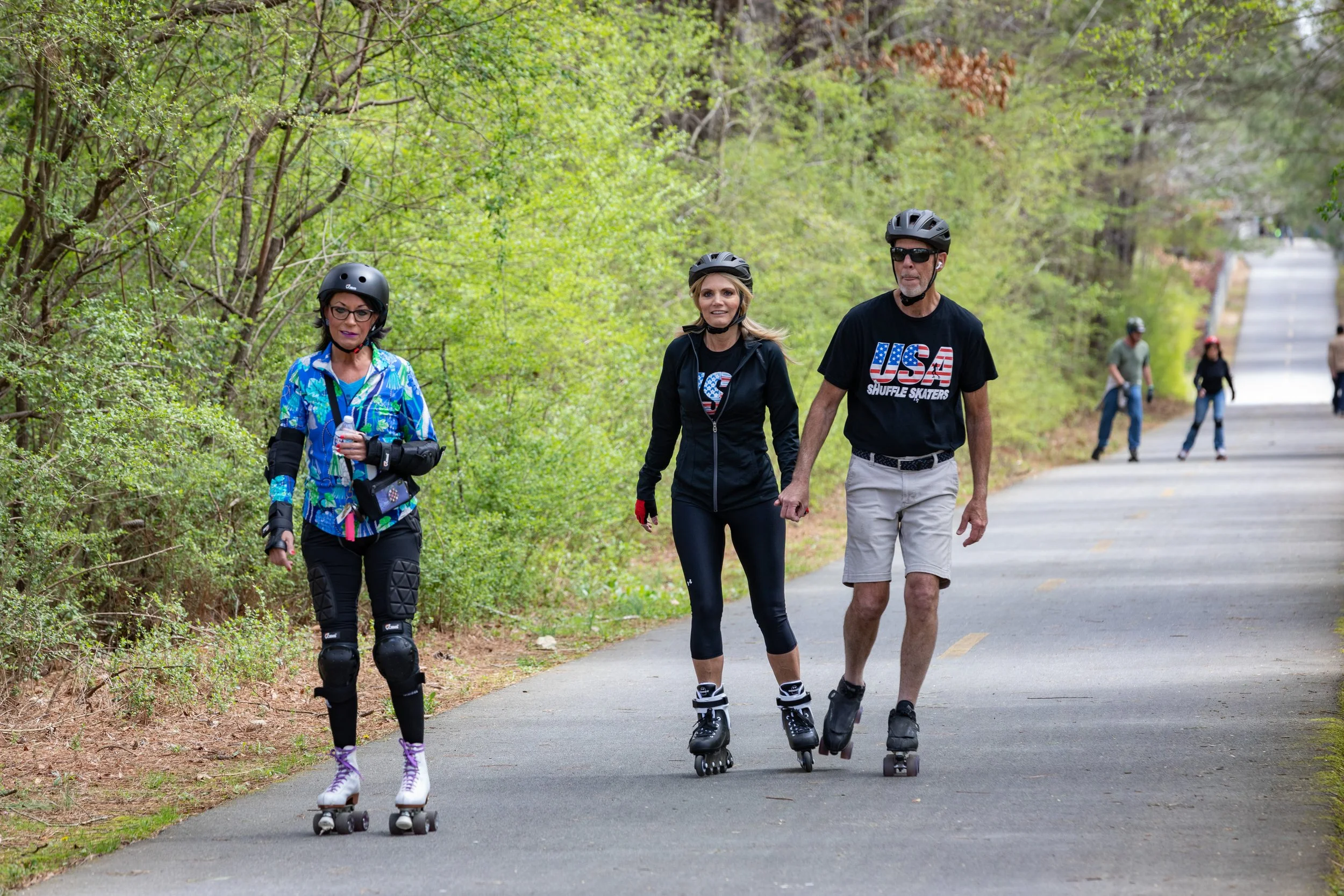 Three people roller skating on a paved trail surrounded by green trees, with others in the background.