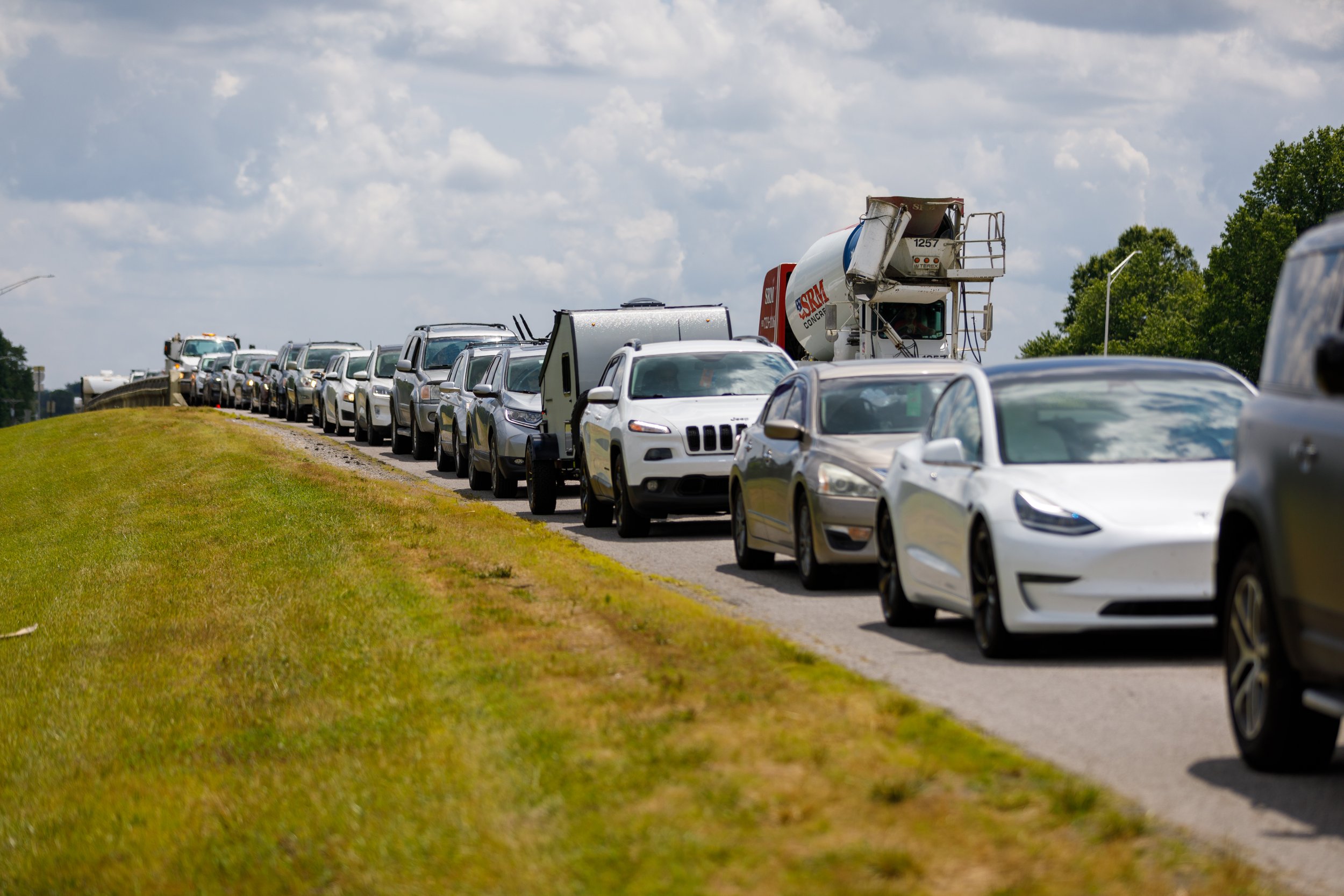 Line of cars on the side of a road under a partly cloudy sky, with some trees in the background.