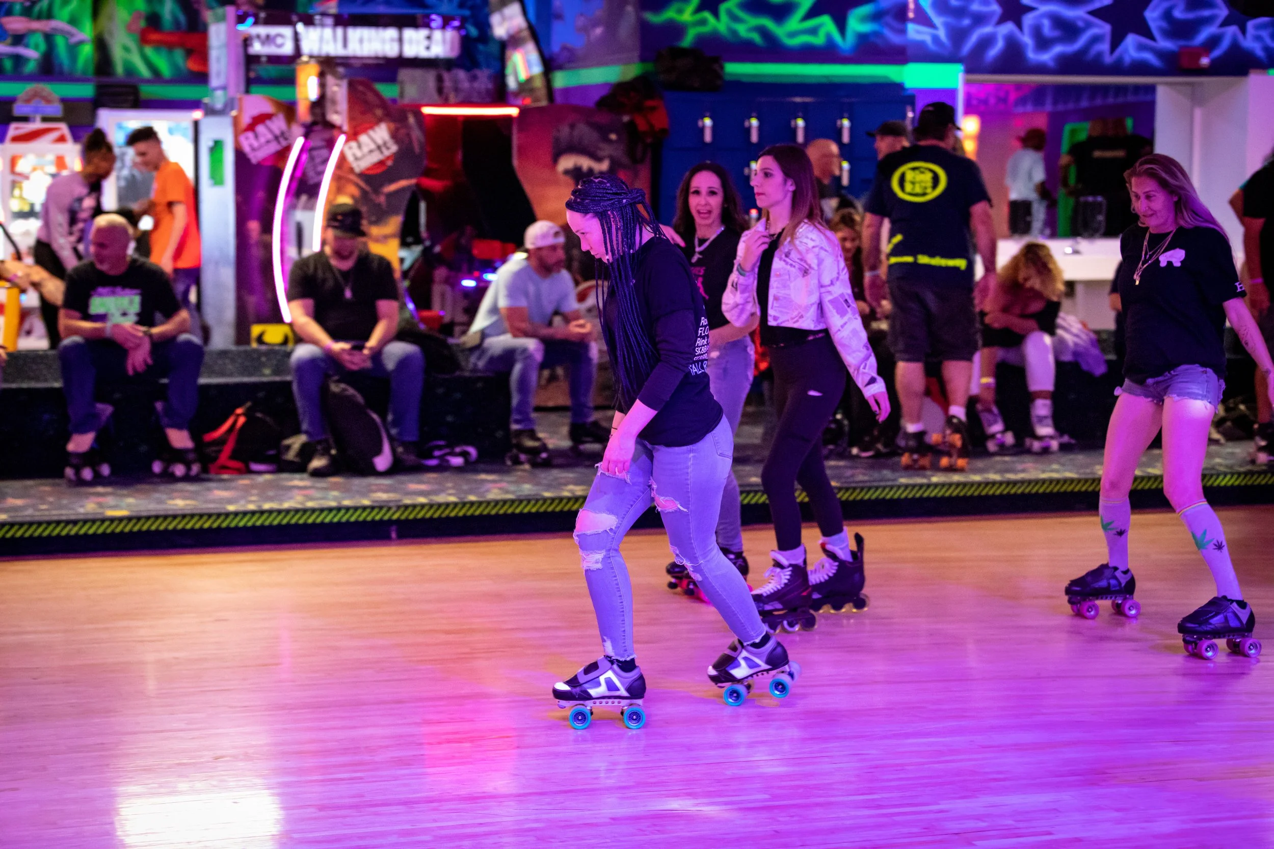 People roller skating indoors at a skate rink with colorful neon lights and wall art, some sitting on benches and others skating.