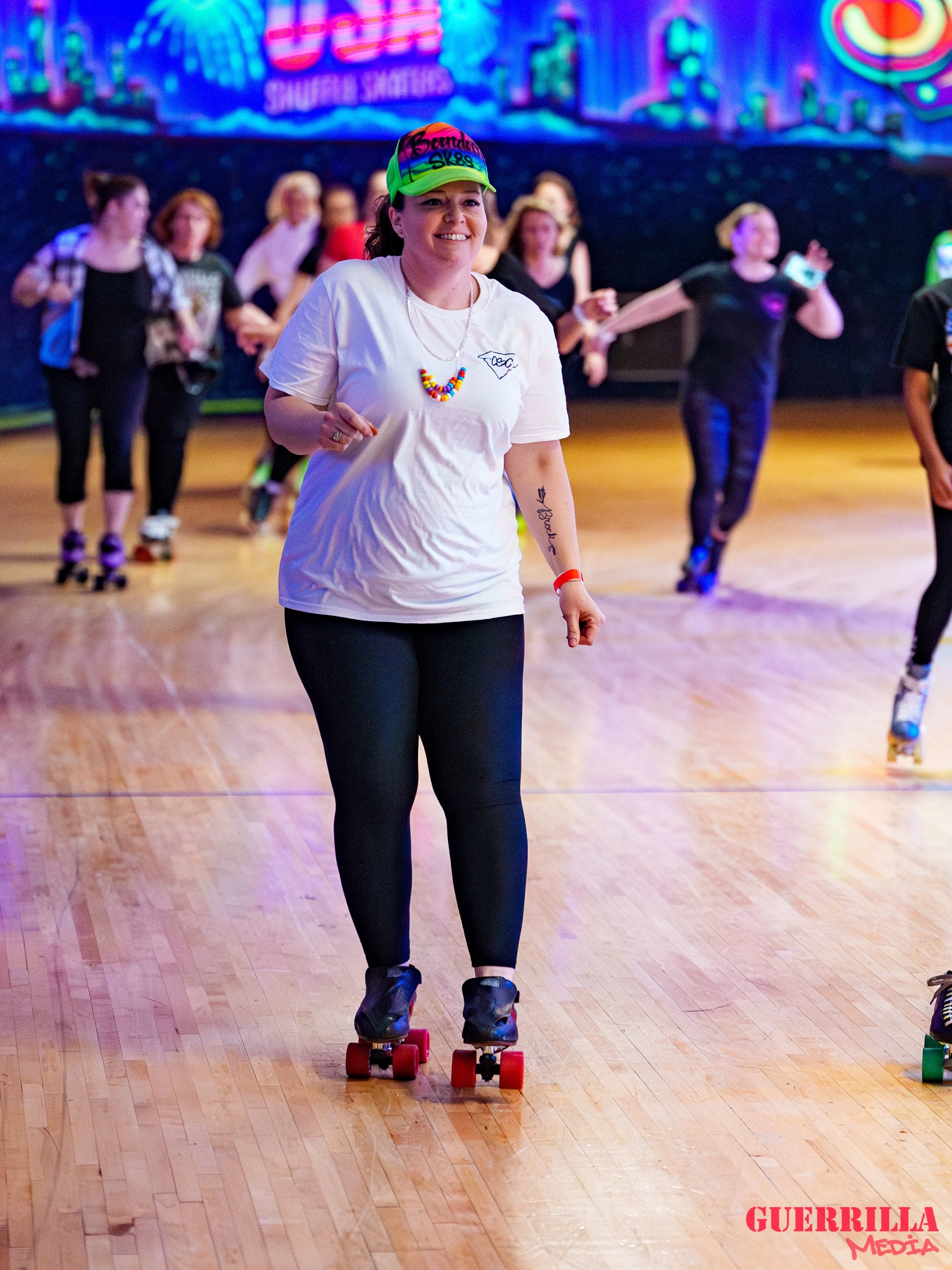 A woman roller skating indoors with a smile on her face, wearing a white t-shirt, black leggings, and a bright colorful cap. She is surrounded by other skaters in the background, with a vibrant, colorful mural on the wall behind them.