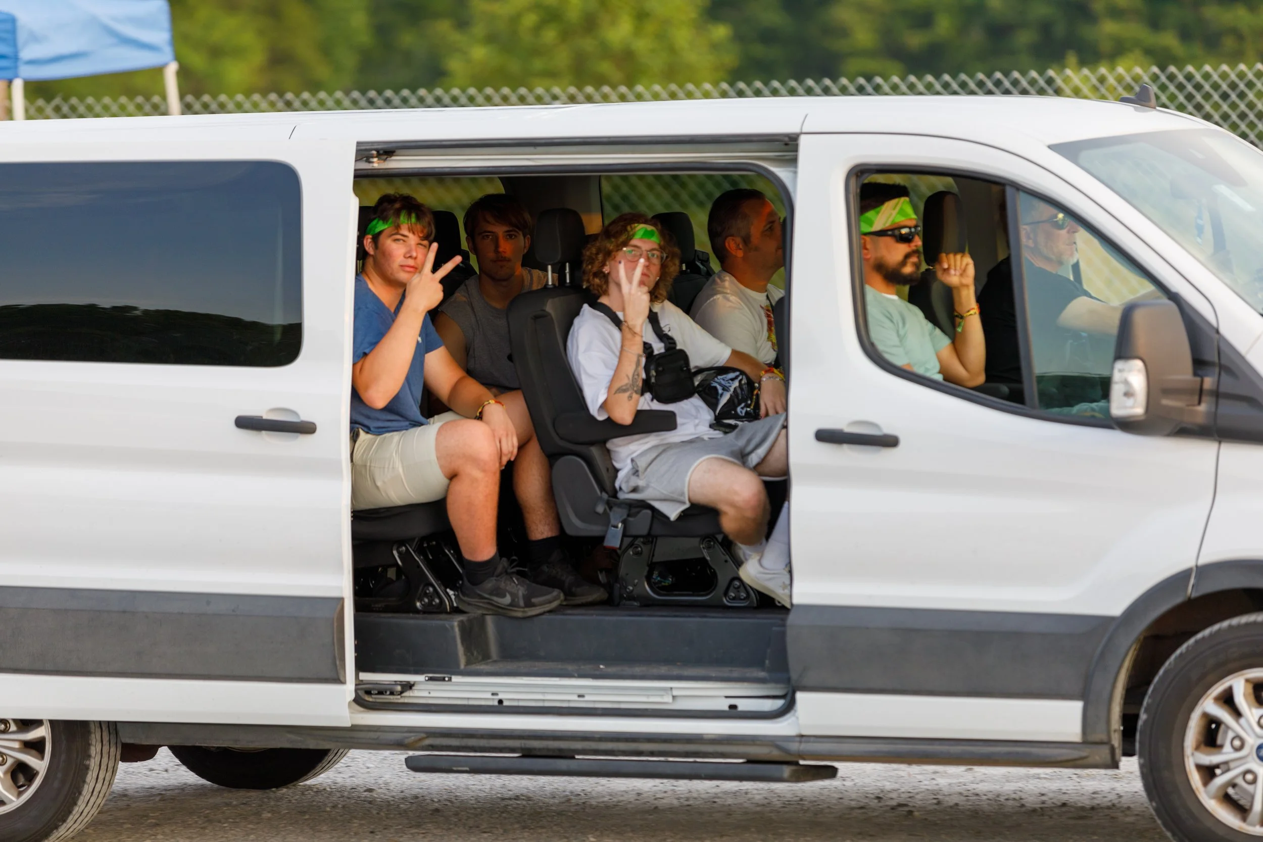 A group of young men seated inside a white van, some making peace signs. They are wearing casual clothes, with two having green headbands and others with sunglasses or glasses. The van has a sliding side door open, revealing the interior and the road