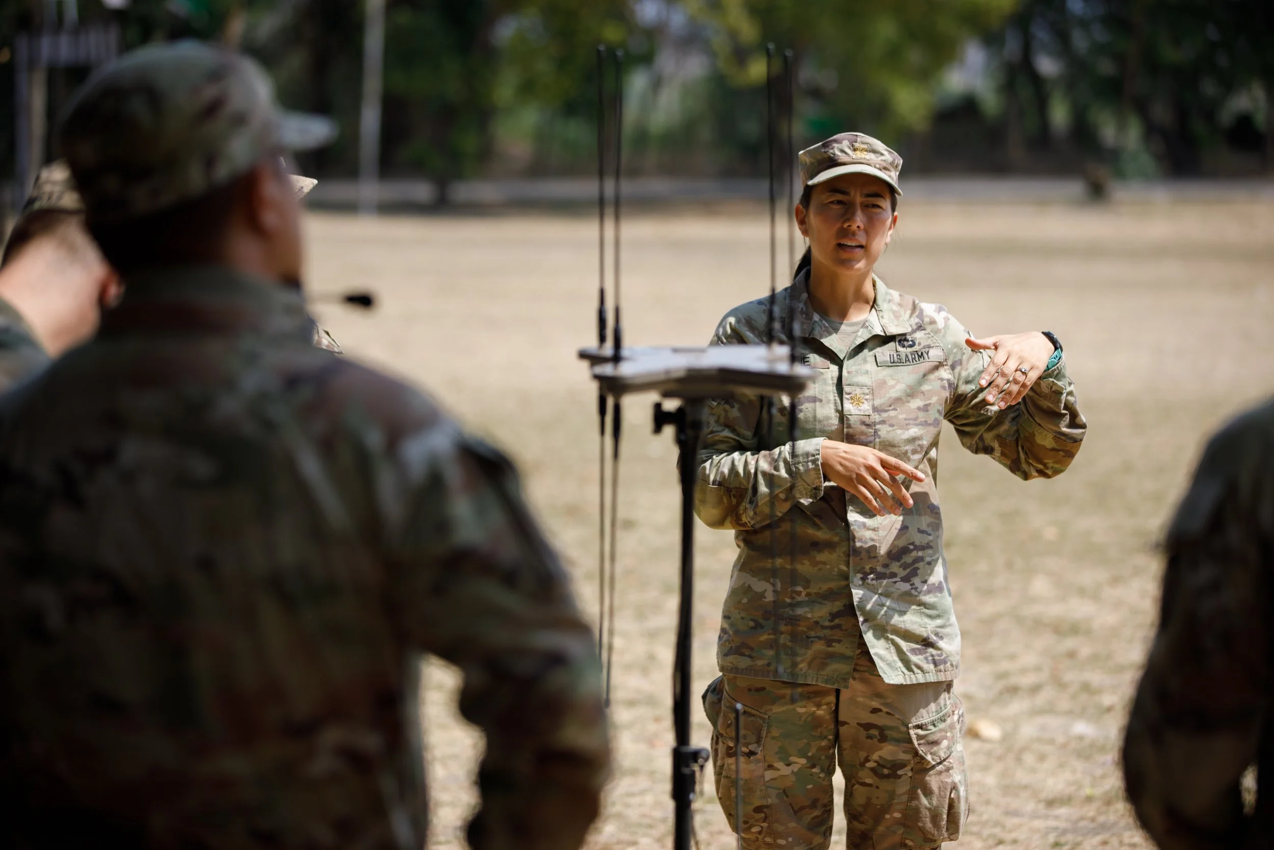 A female U.S. Army soldier in camouflage uniform speaking and gesturing while standing outdoors, with other soldiers in similar uniforms listening.