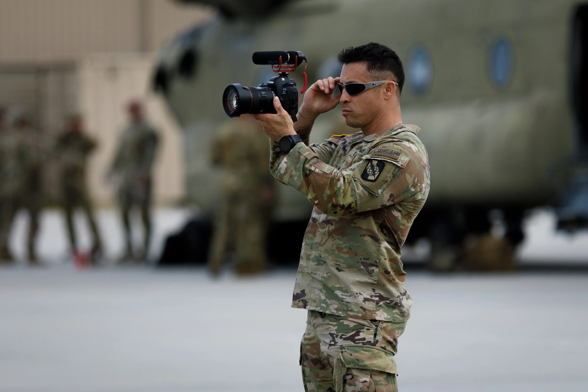 Military man in camouflage uniform filming with a camera, with soldiers and a helicopter in the background.