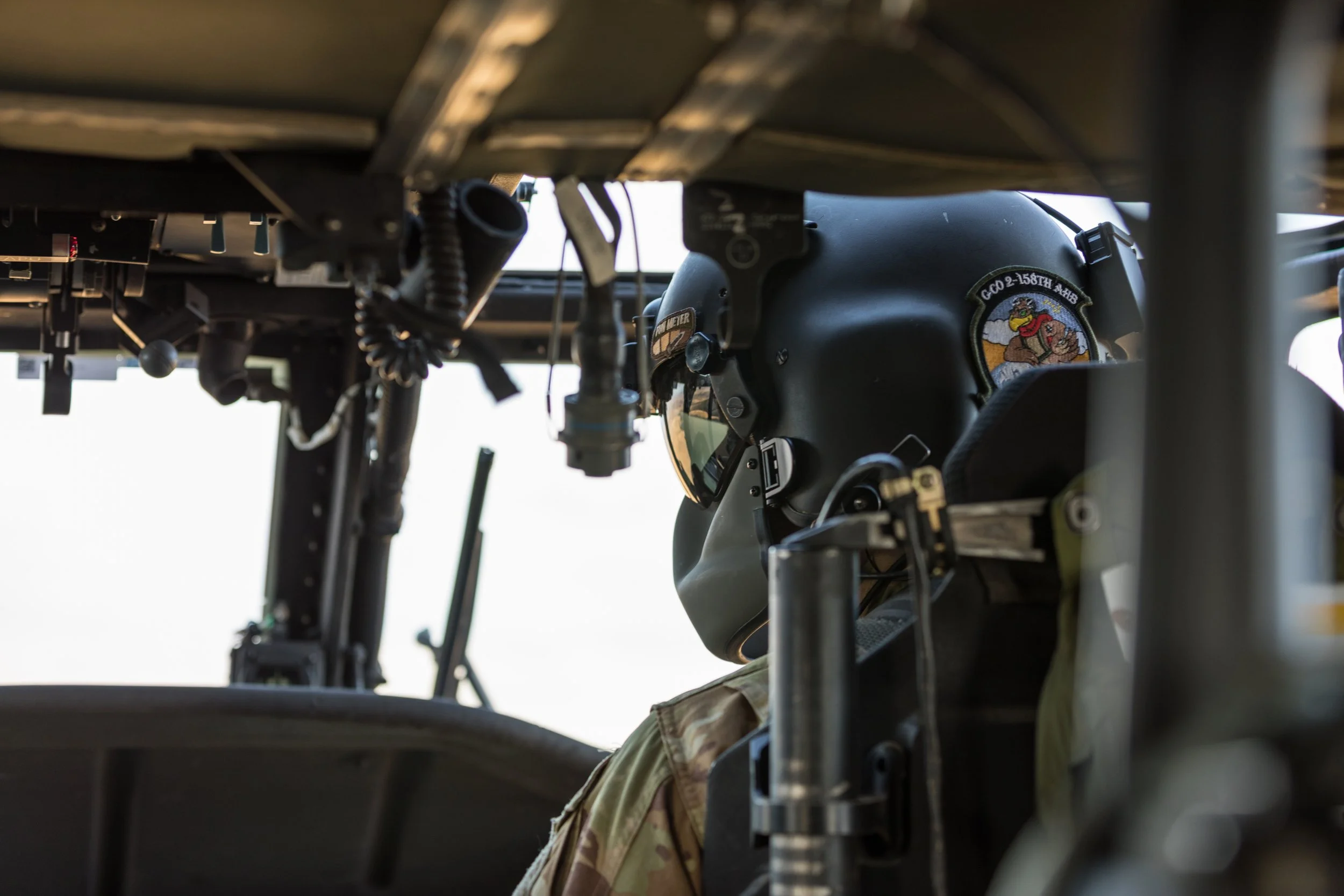 Close-up of a military pilot wearing a black helmet with a patch, sitting inside an aircraft cockpit with visible controls and equipment.