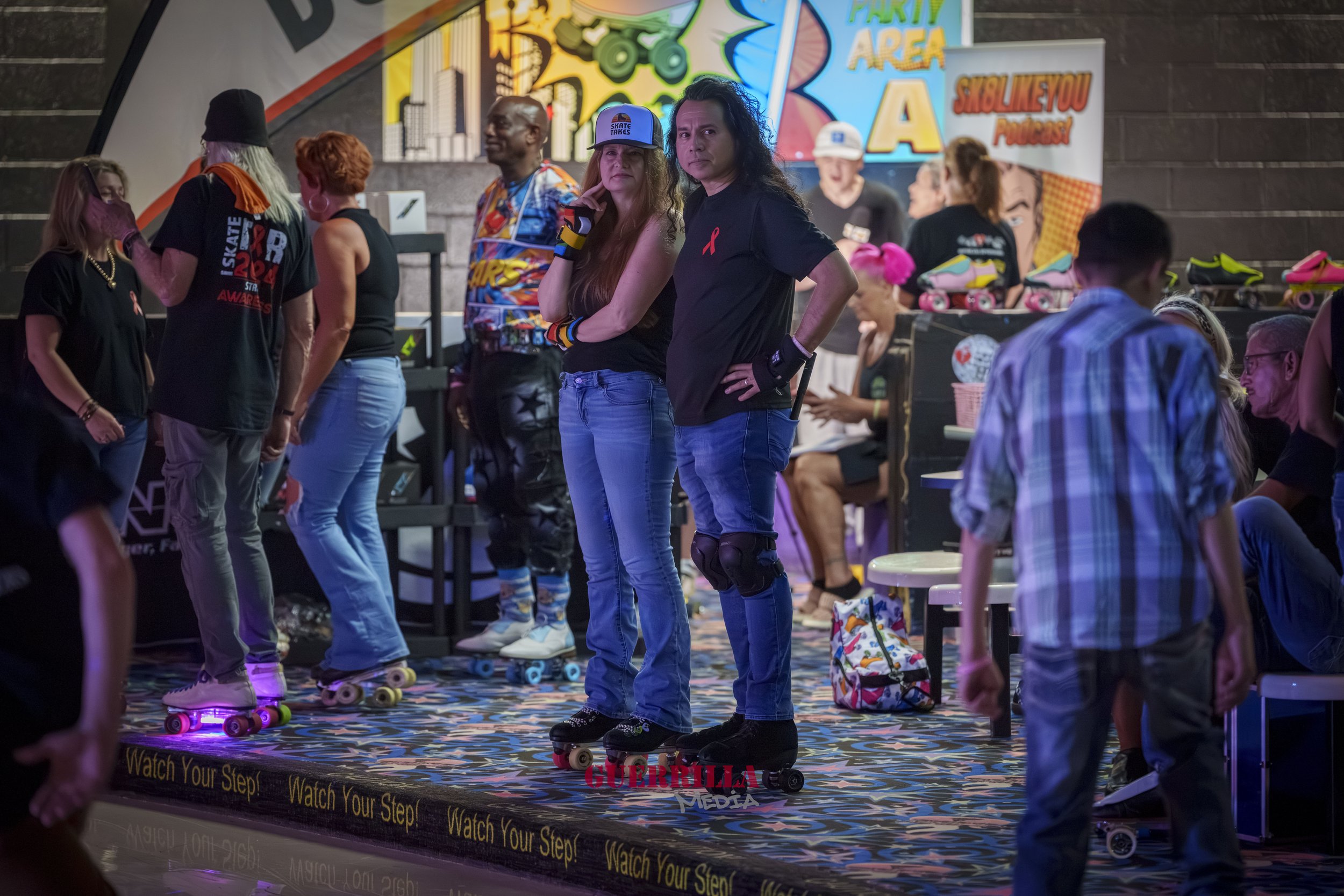 People roller skating in an indoor rink. Several individuals are standing or skating, some engaged in conversation. Brightly colored decor and banners are visible in the background, including a sign that reads 'PARTY AREA.' The floor has a colorful, 
