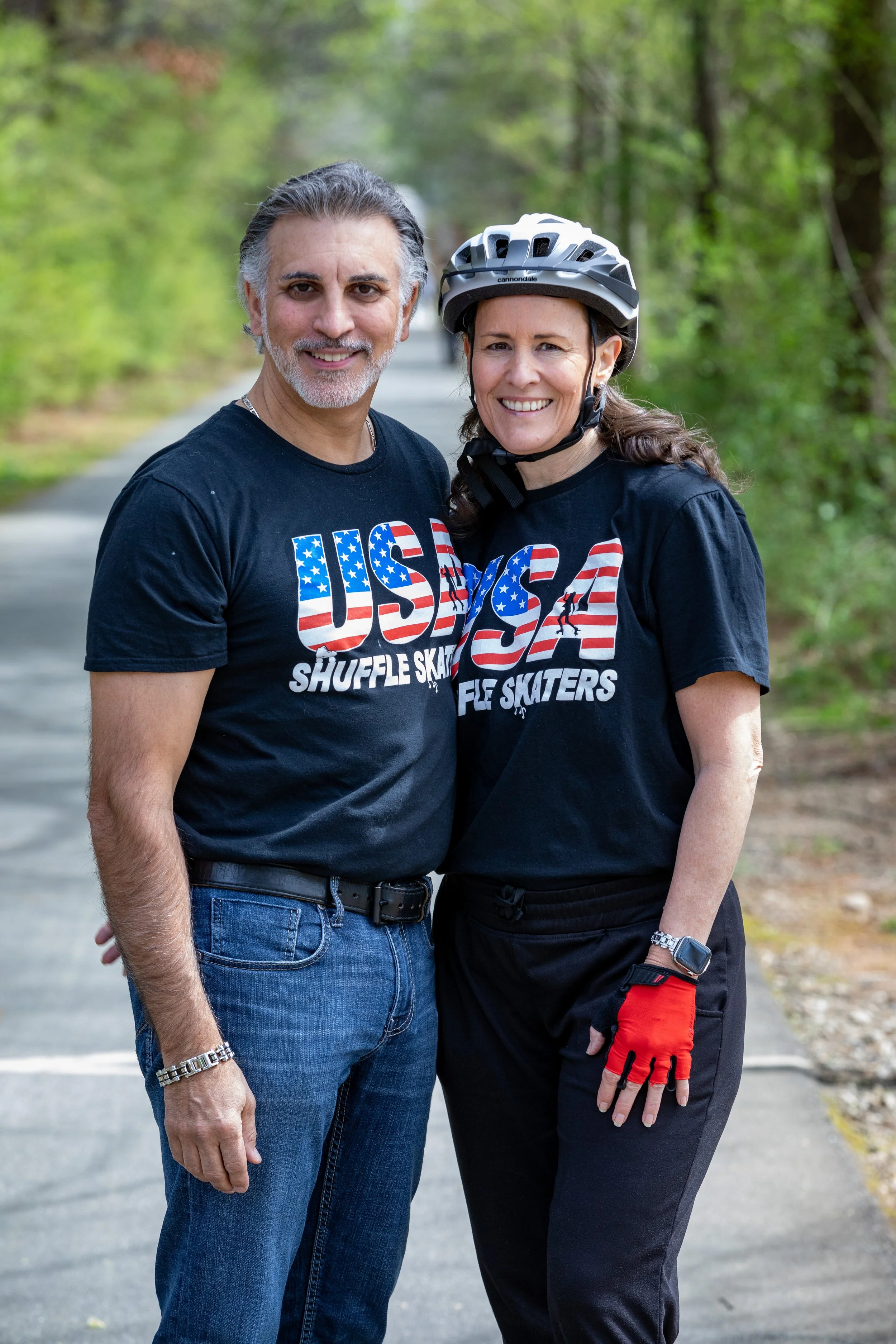 A smiling middle-aged man and woman stand outdoors on a paved path surrounded by green trees. They are wearing matching black T-shirts with an American flag-themed "USA Shuffle Skaters" logo. The woman is wearing a bicycle helmet and red gloves.