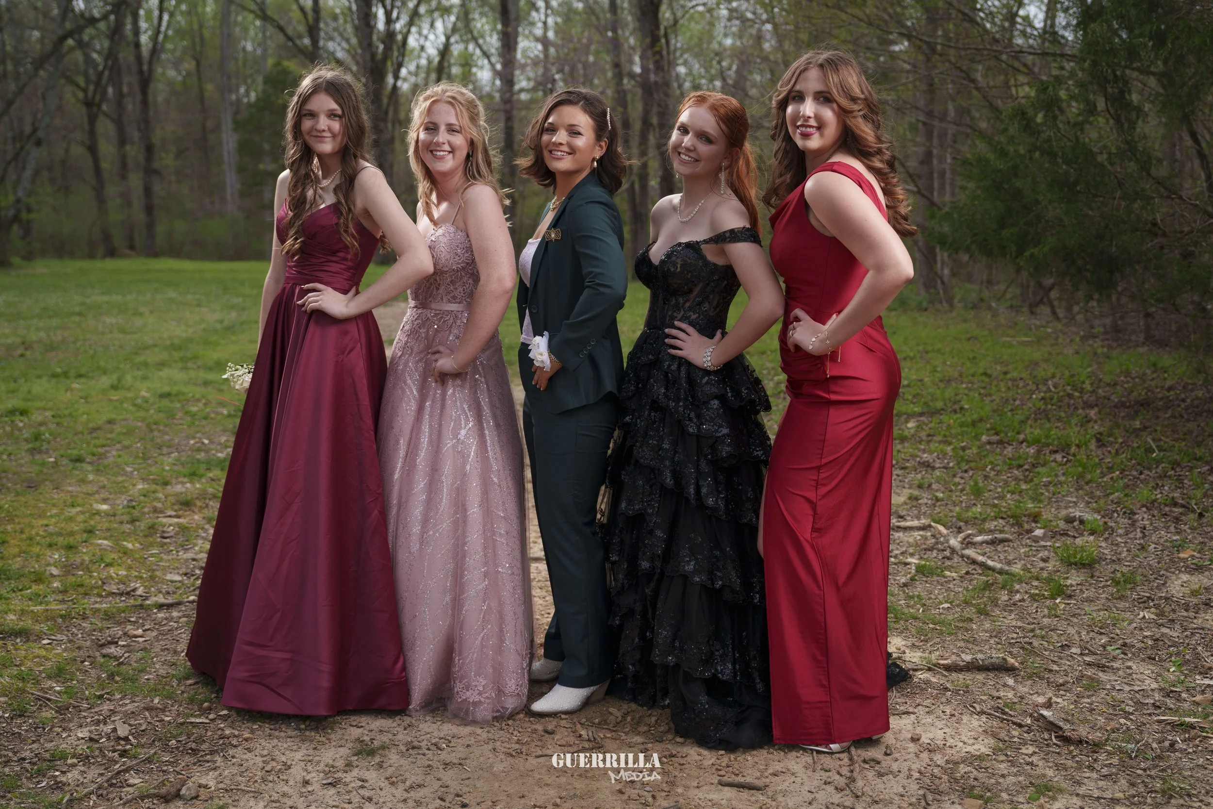 Five young women in formal dresses standing outdoors on a dirt path in a wooded area, smiling and posing for a photo.