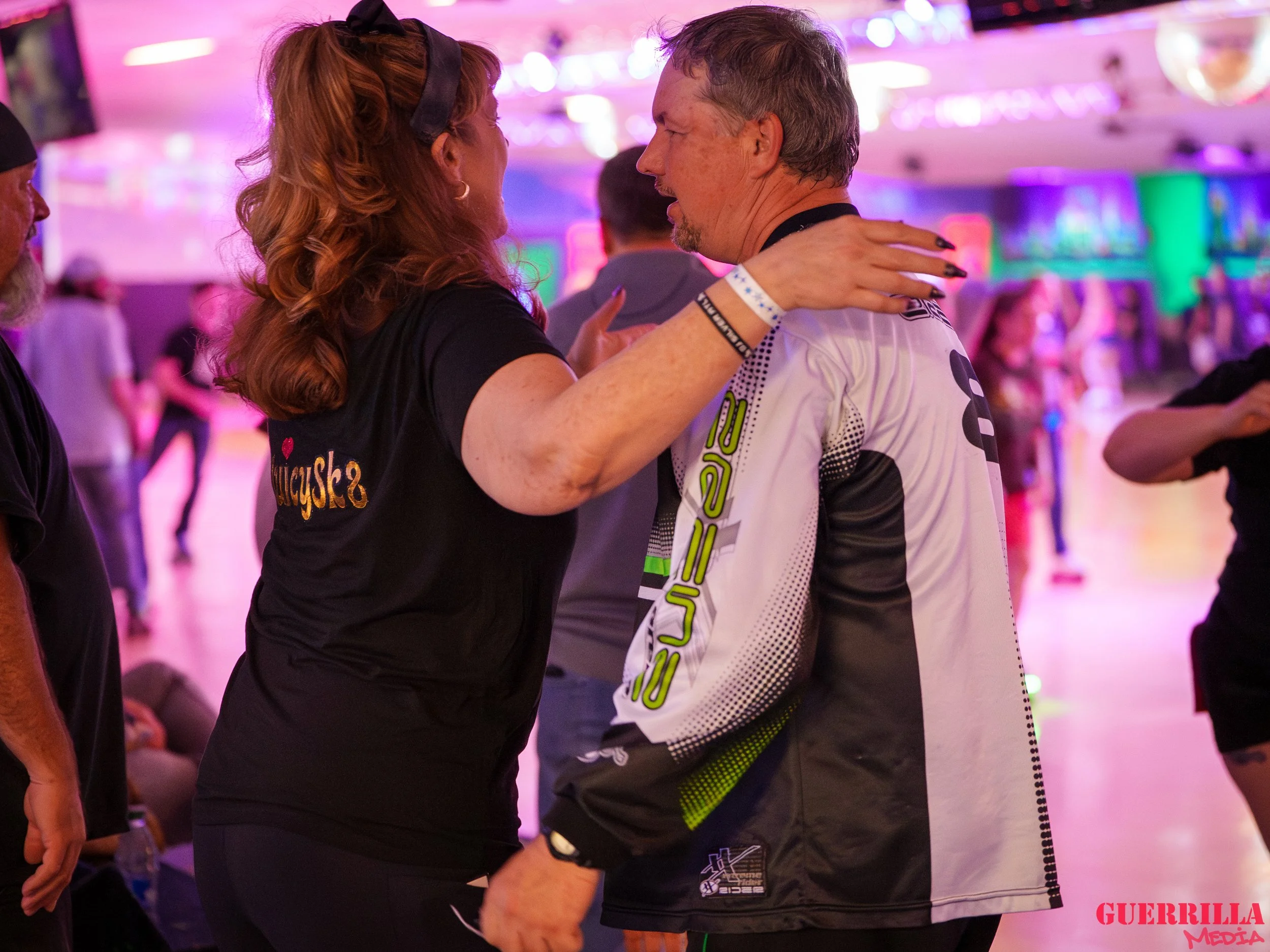 A woman with red hair and a black t-shirt is embracing and talking to a man in a white and black sports jacket; they are inside a roller-skating rink with colorful lighting and other skaters in the background.
