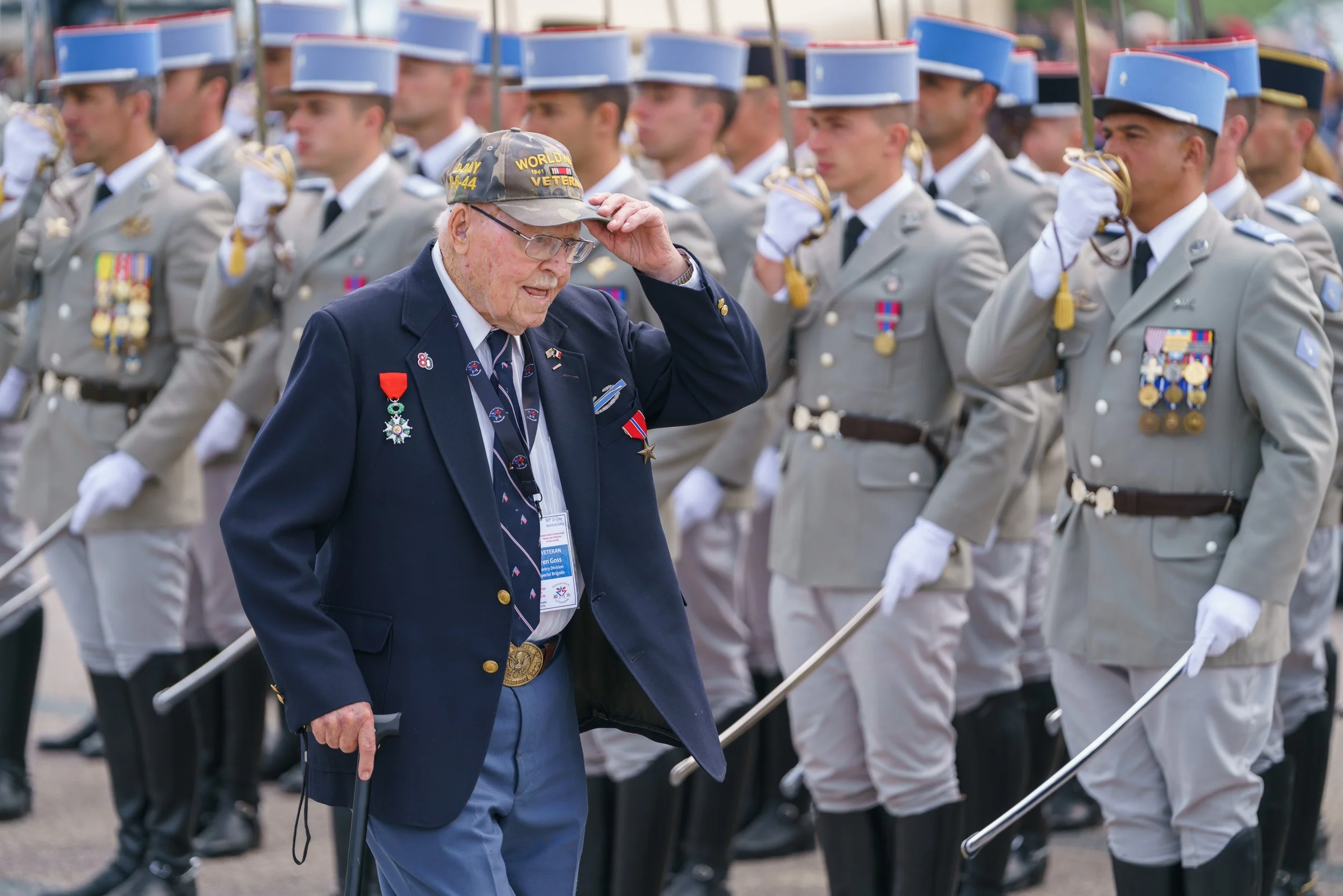 Elderly veteran saluting during a military parade, with soldiers in uniform standing in formation behind him.
