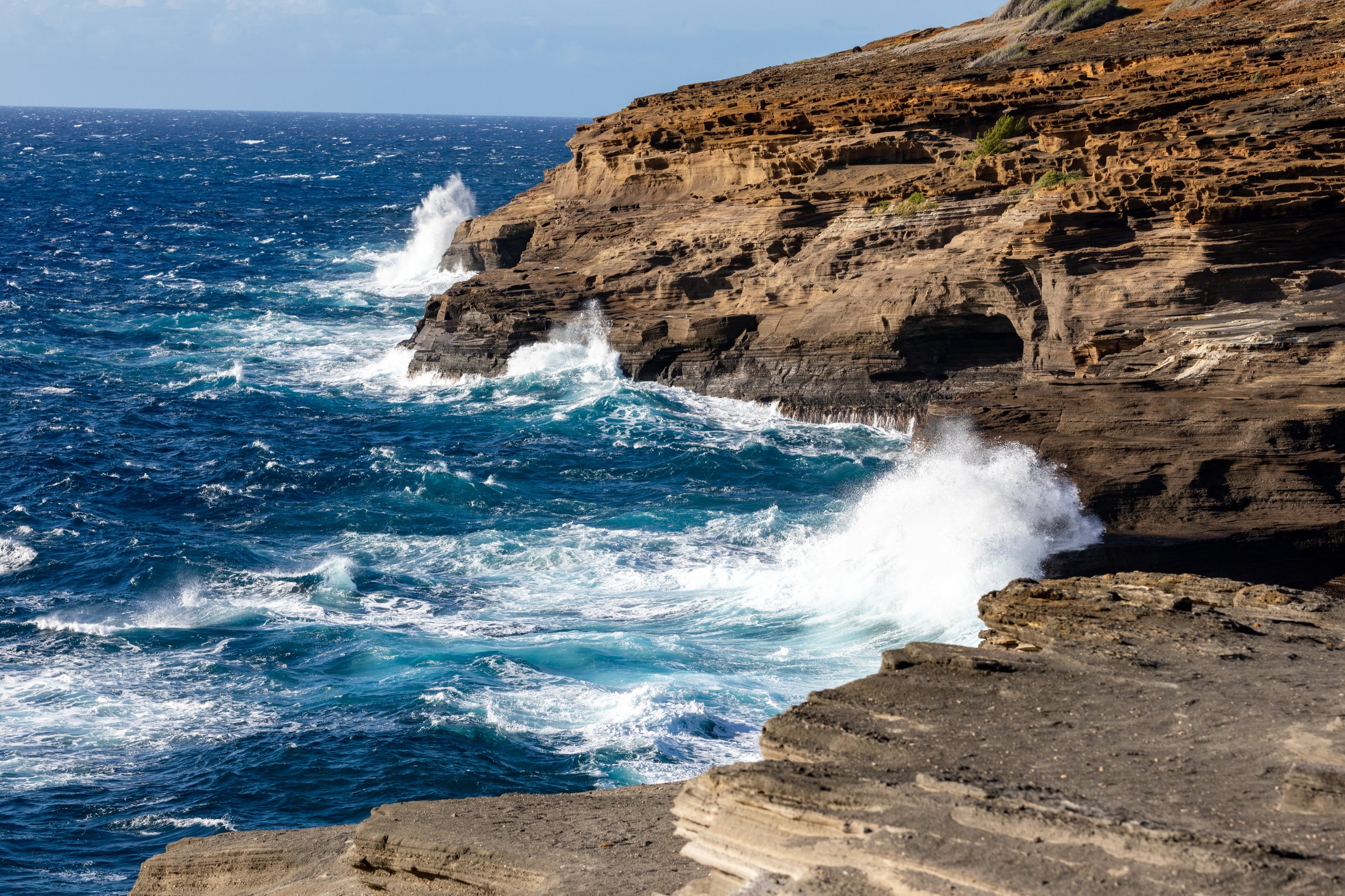 The image shows ocean waves crashing against rocky cliffs and shoreline under a partly cloudy sky.