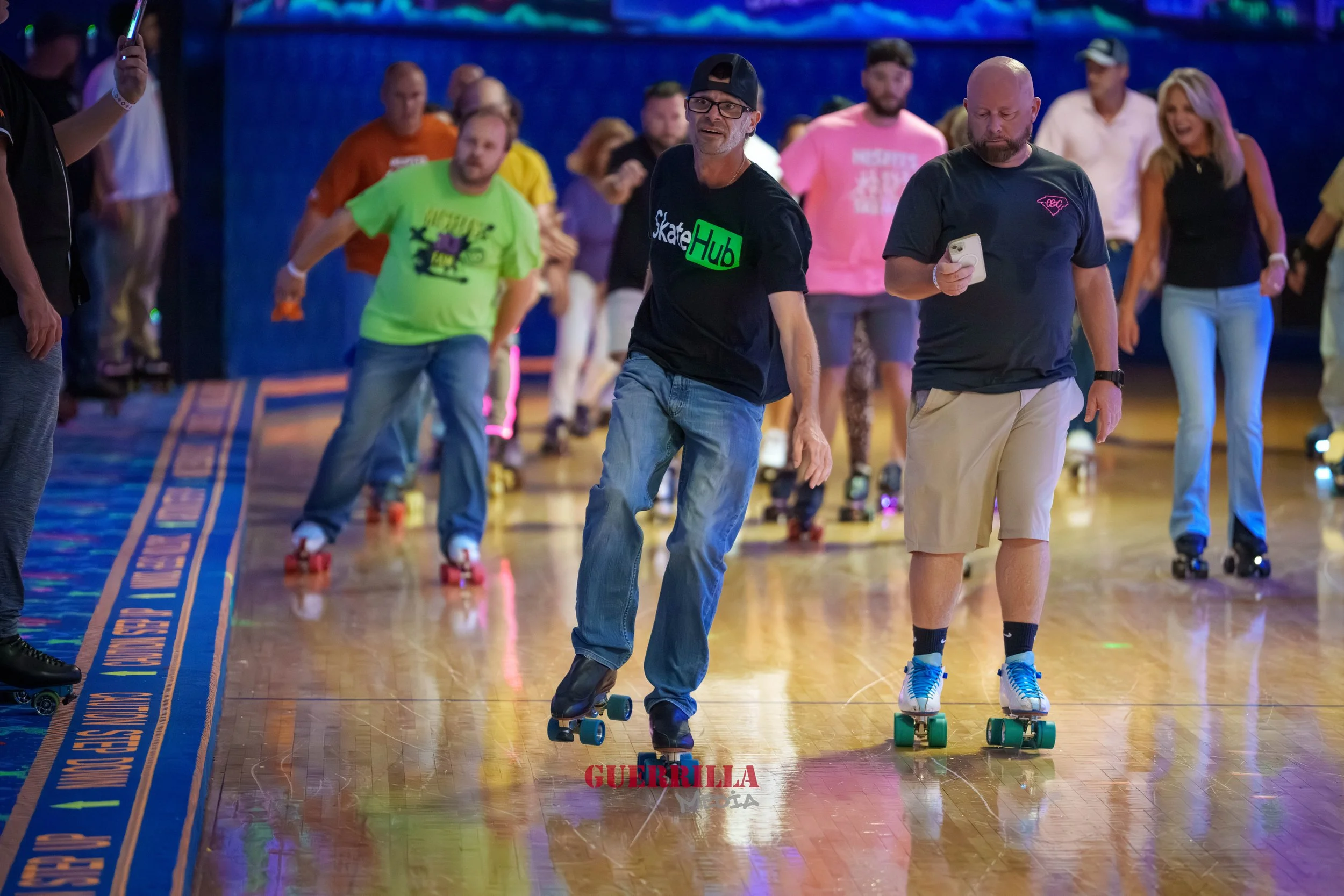 People roller skating indoors, with a man wearing a black t-shirt that says 'Skate Hub' and another man holding a phone watching the skaters.