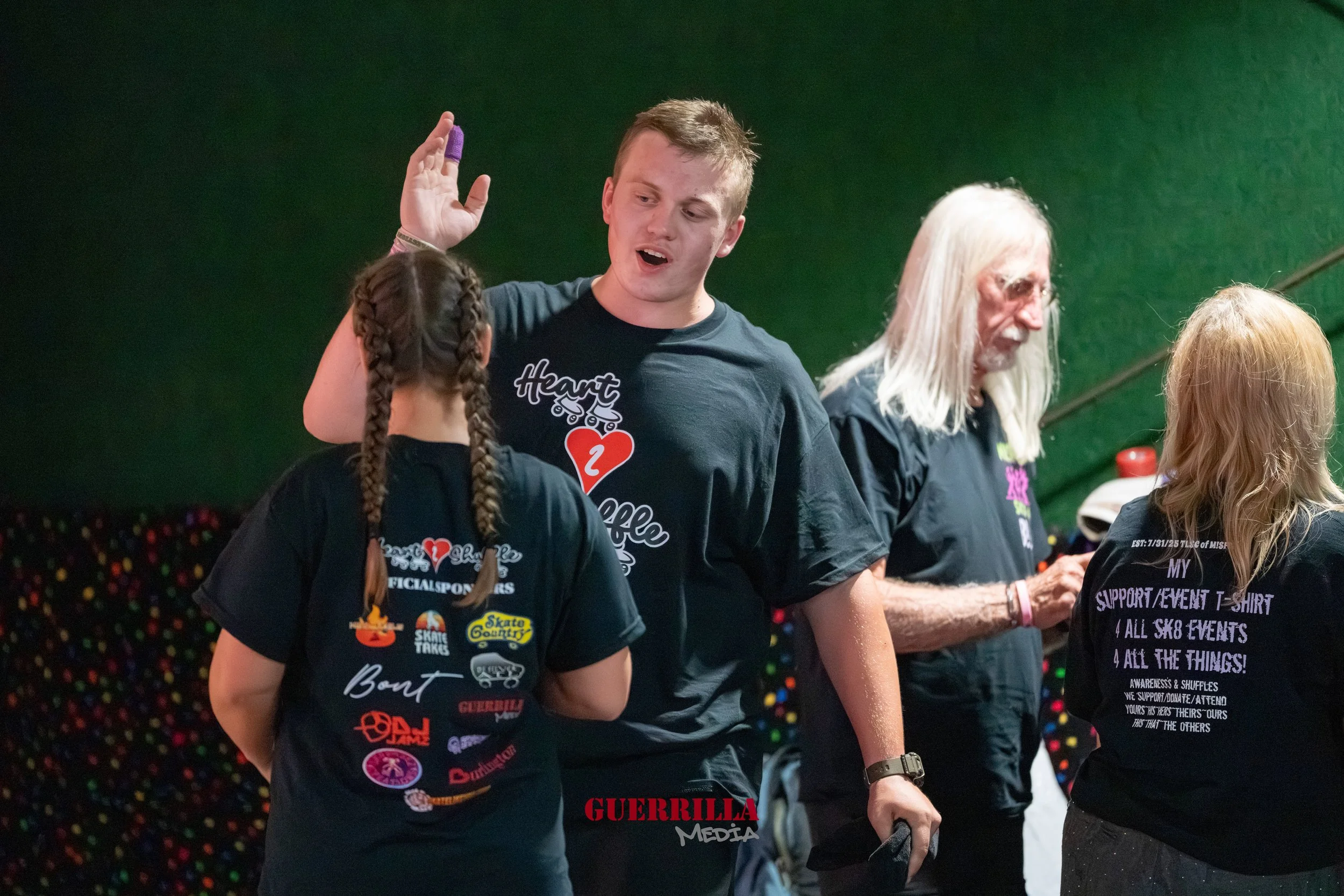 Group of people at a skating rink, engaging in conversation, with a green wall in the background and colorful dots on the lower part of the wall.