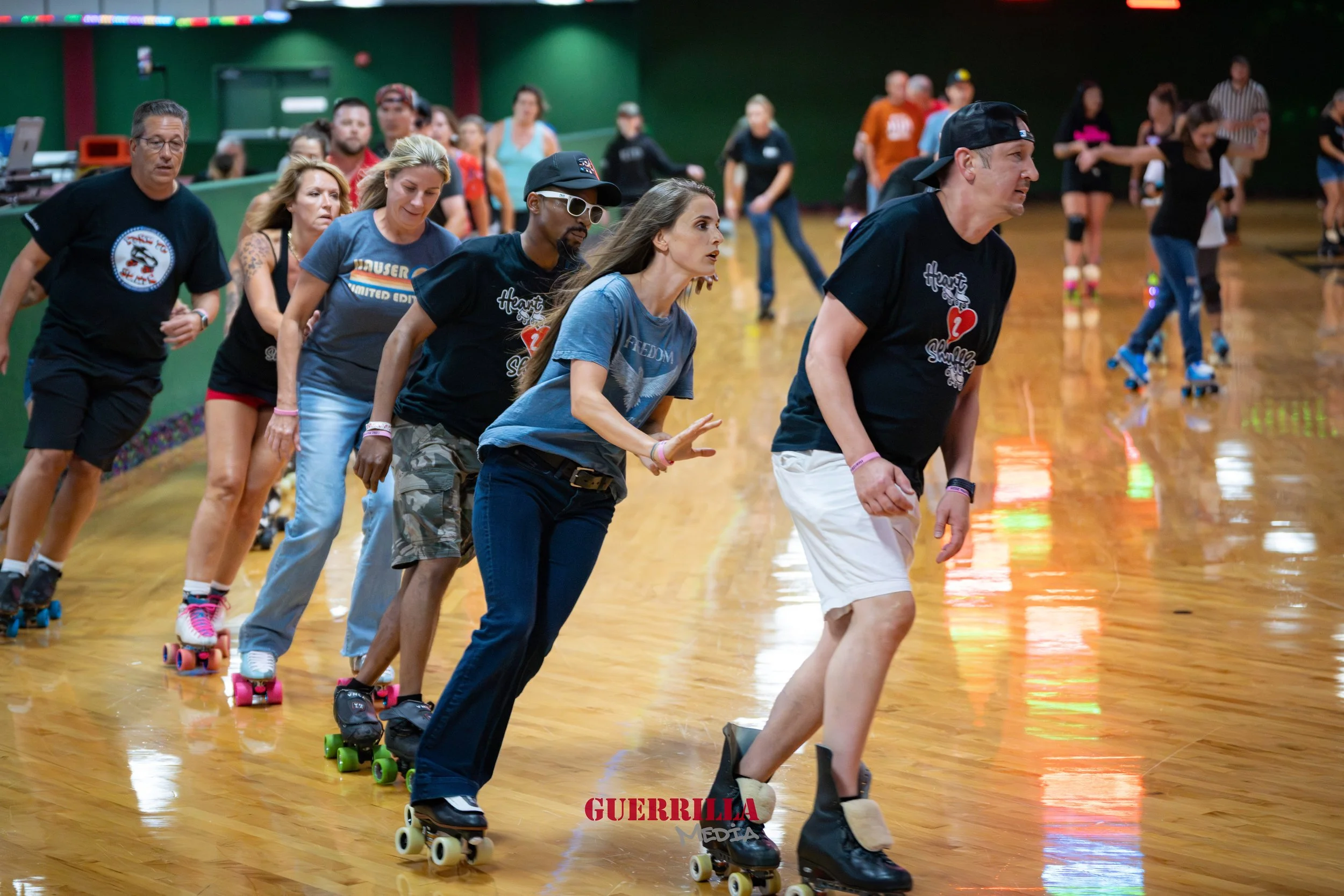 A group of people roller skating together indoors, with some of them in the background and others in the foreground, on a wooden rink.