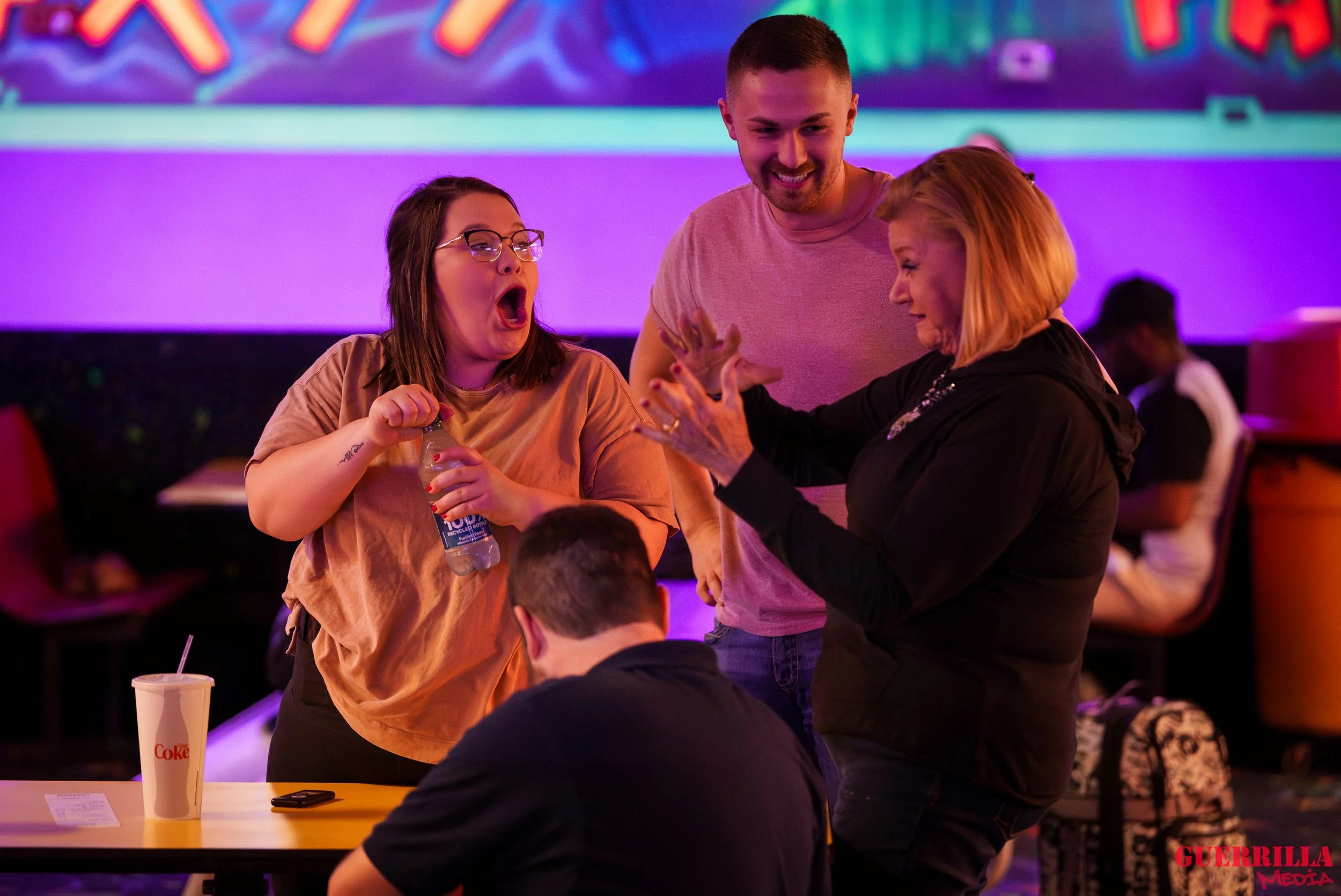 A group of four people in a lively setting. Two women and two men. The woman on the left is holding a water bottle and appears to be yelling or laughing with her mouth open. The woman on the right has blonde hair and is gesturing with her hands. One 