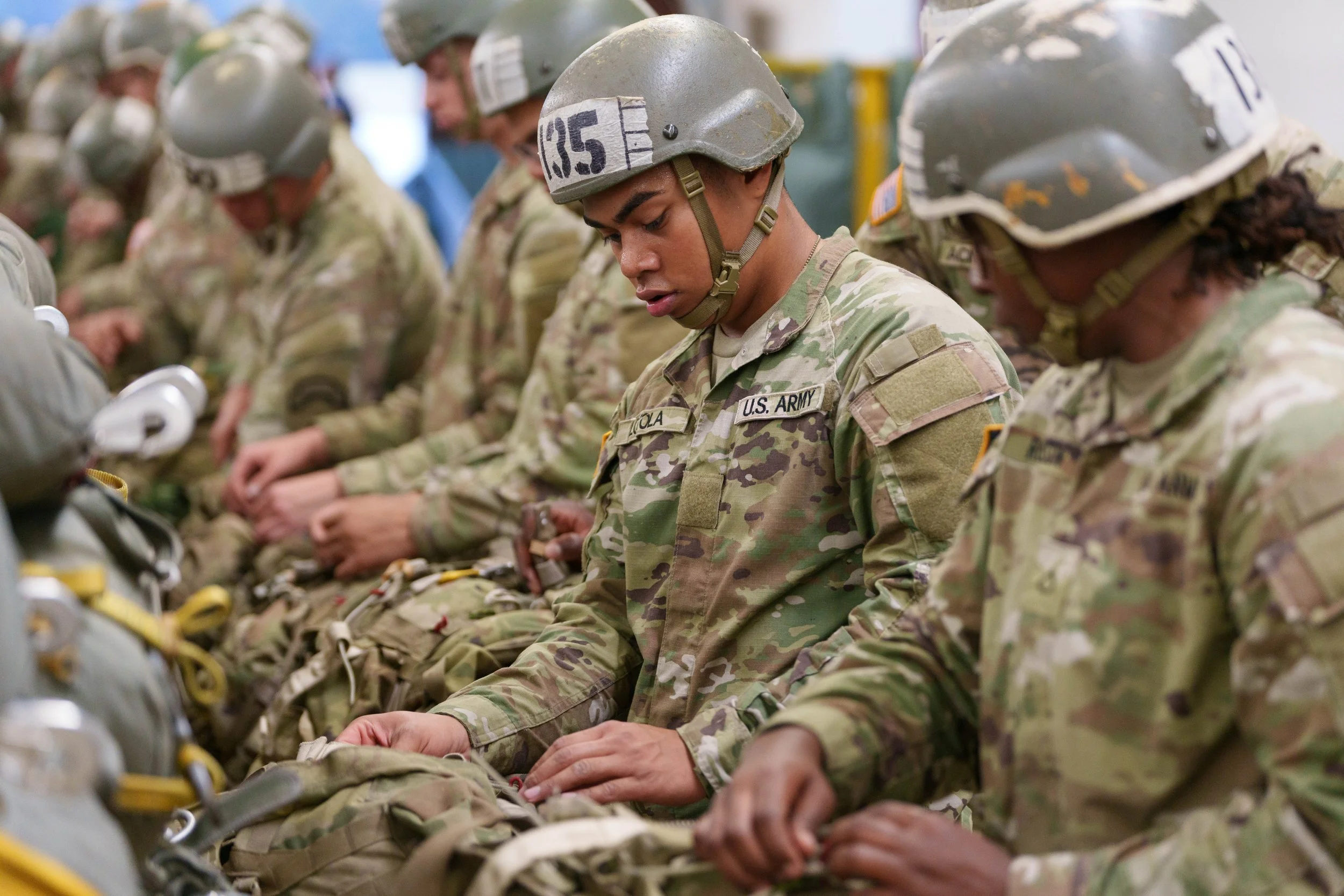 U.S. Army soldiers in camouflage uniforms and helmets sitting in a row, preparing their gear.