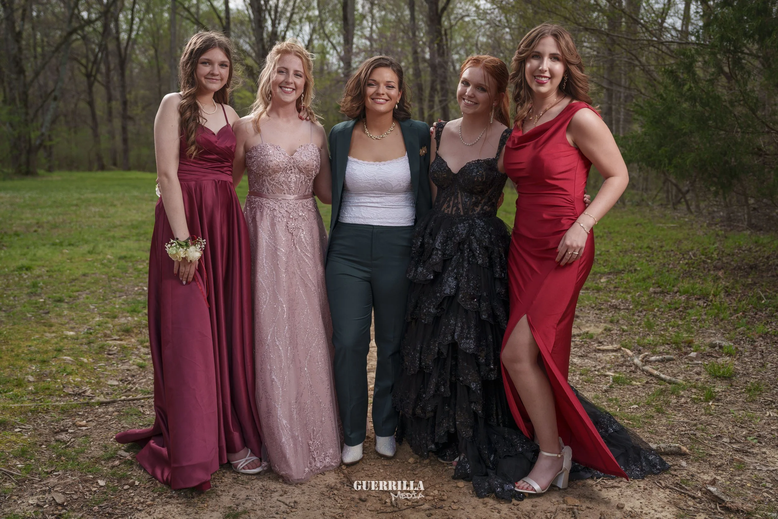 Five young women dressed in formal gowns and a woman in a suit are standing outdoors on a grassy area with trees in the background, smiling for a group photo.