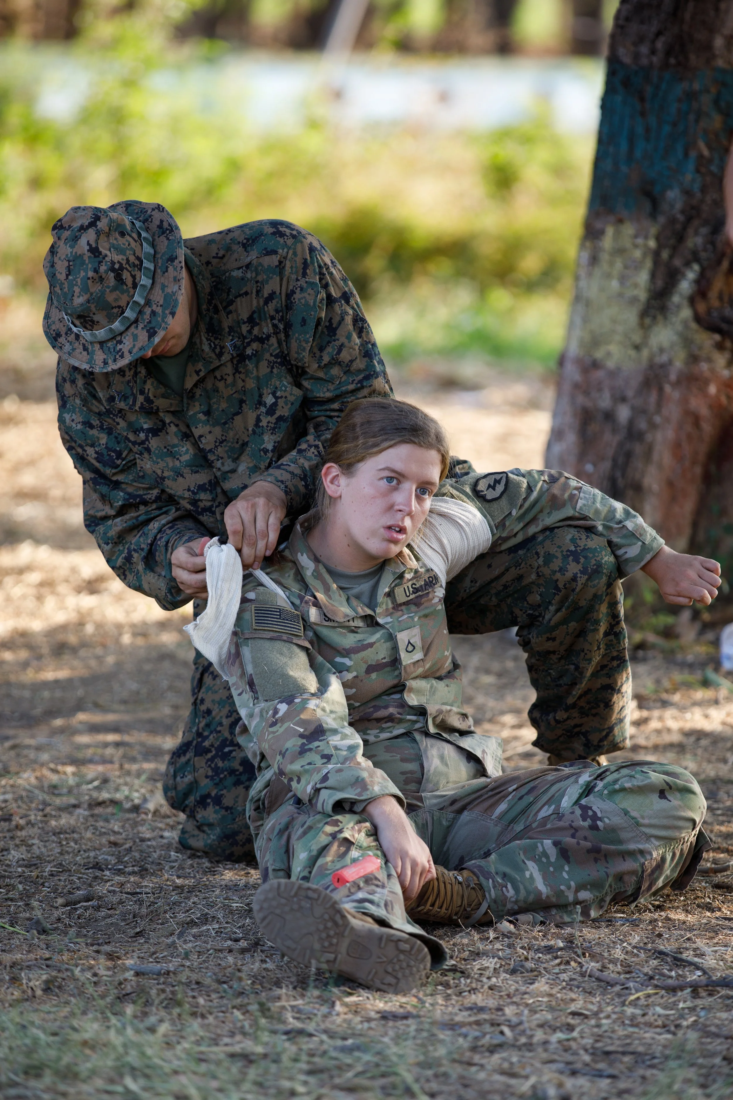 A female soldier sitting on the ground outdoors with a distressed expression, being attended to by an individual in military camouflage uniform, possibly a medic, who is assisting her after an injury.