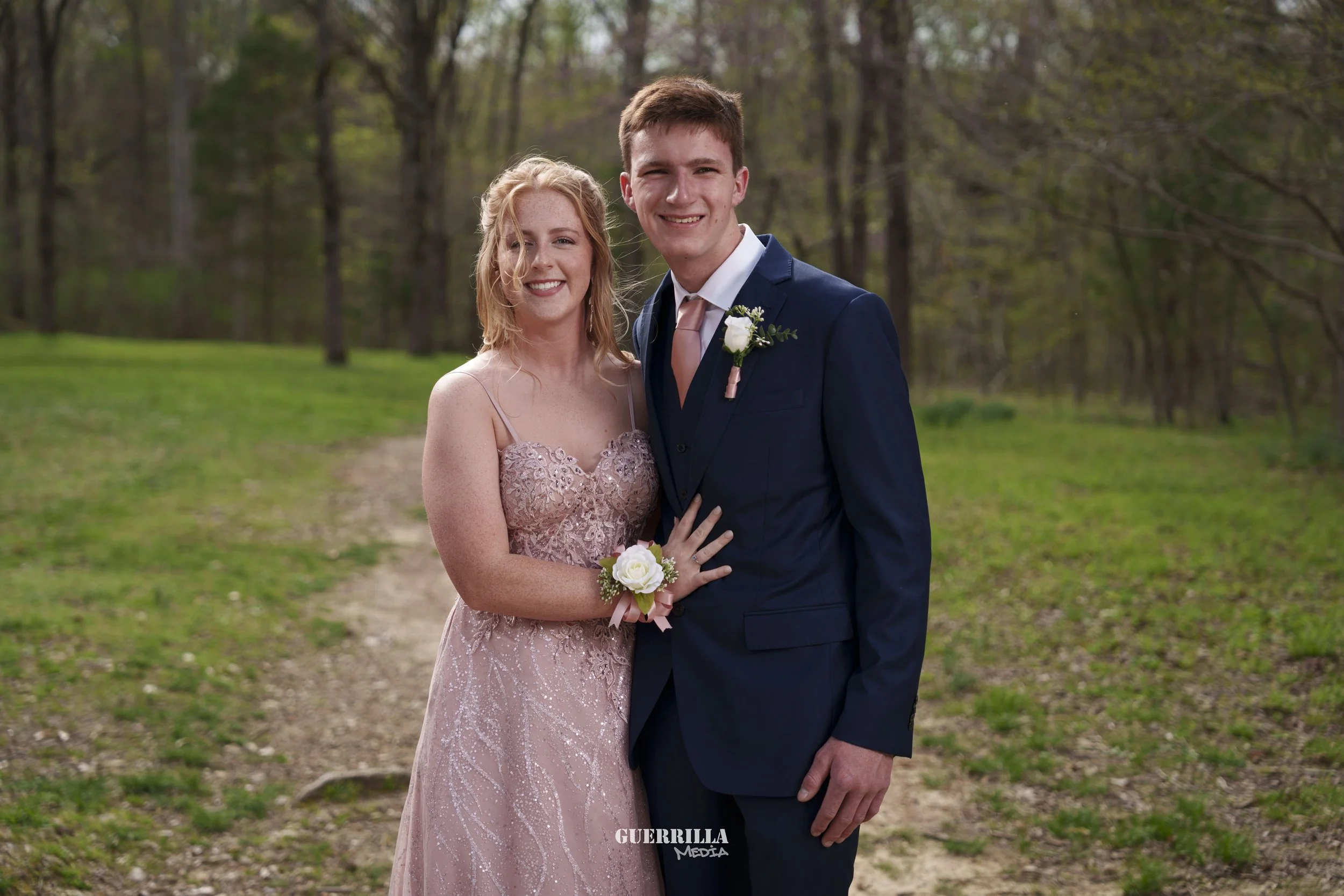 A young couple in formal attire standing on a forest path, smiling at the camera, with the woman wearing a pink lace dress and the man in a navy suit with a boutonniere, during daytime.