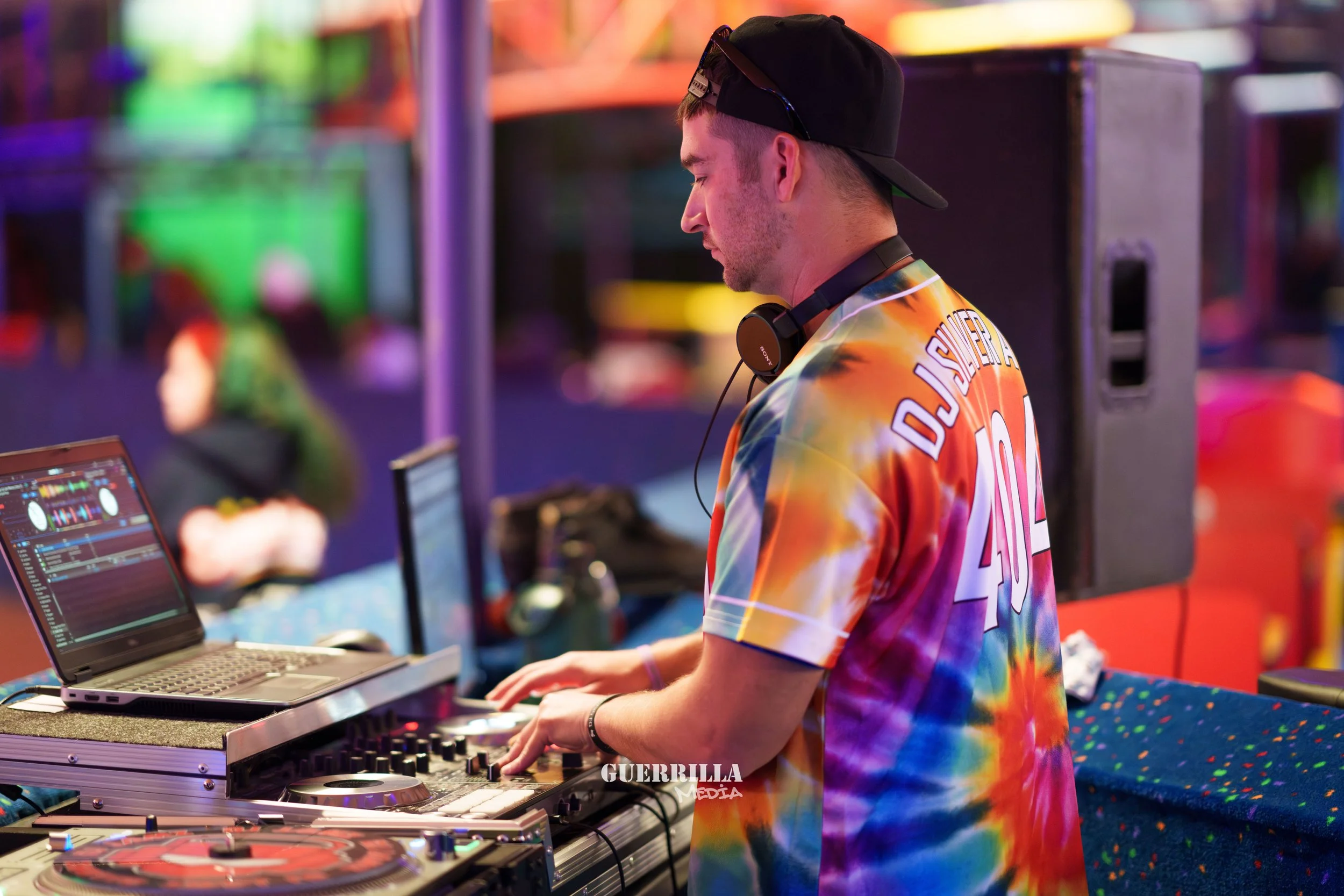 A DJ wearing a black cap and a colorful tie-dye shirt with the number 40 on the back, standing at a DJ booth, mixing music with headphones around his neck, in a vibrant, colorful nightclub setting.