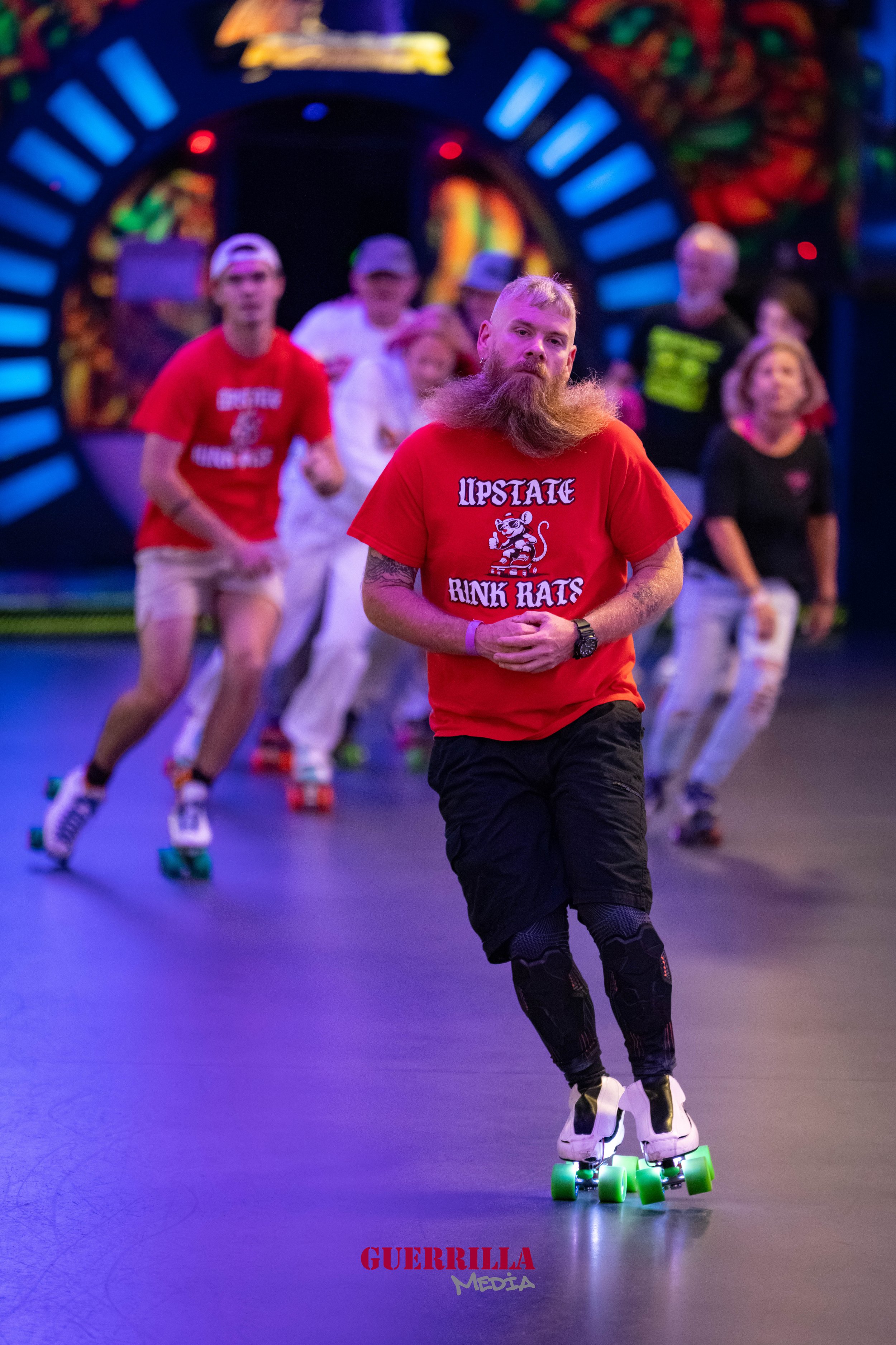 A group of people roller skating indoors, with a bearded man in the foreground wearing a red t-shirt that says 'UPSTATE RINK RATS', and others in the background also skating, under colorful neon lights.