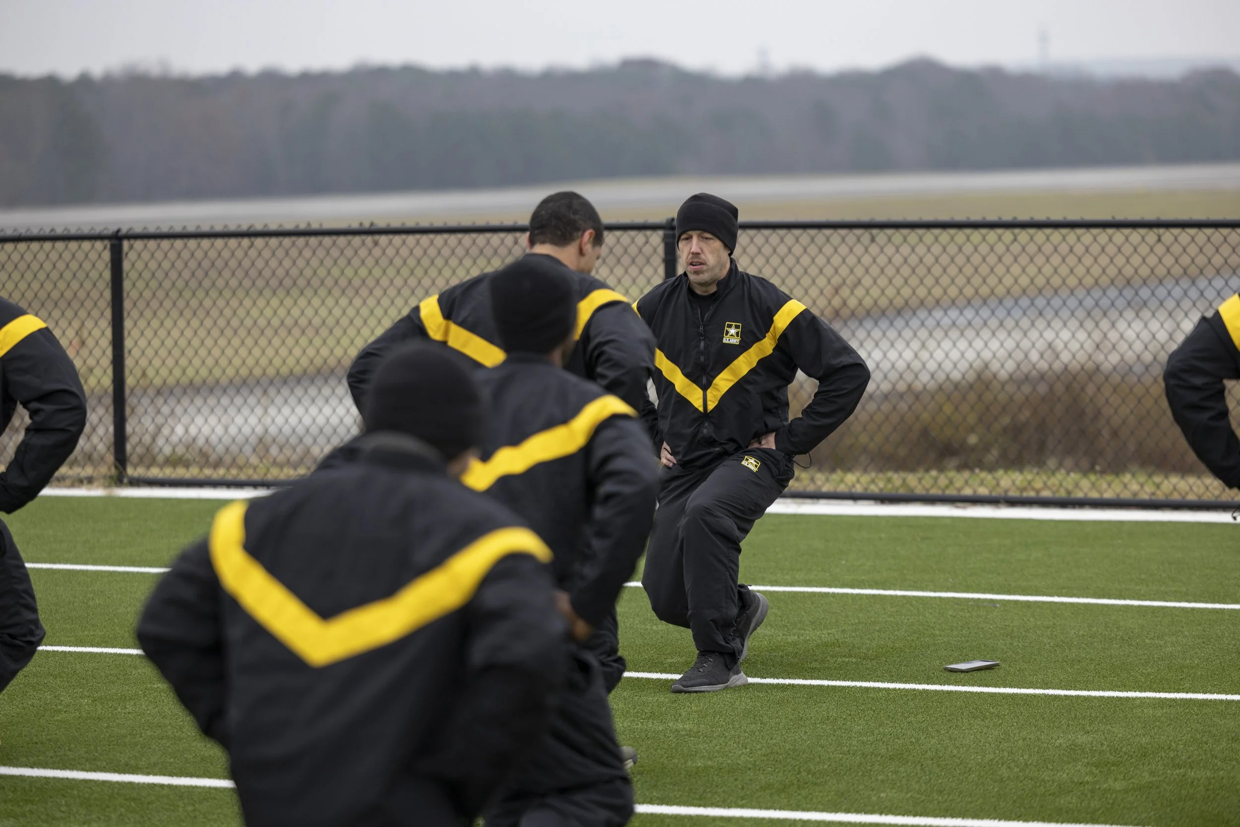 Group of athletes in black and yellow training on a football field outside, with a coach giving instructions.