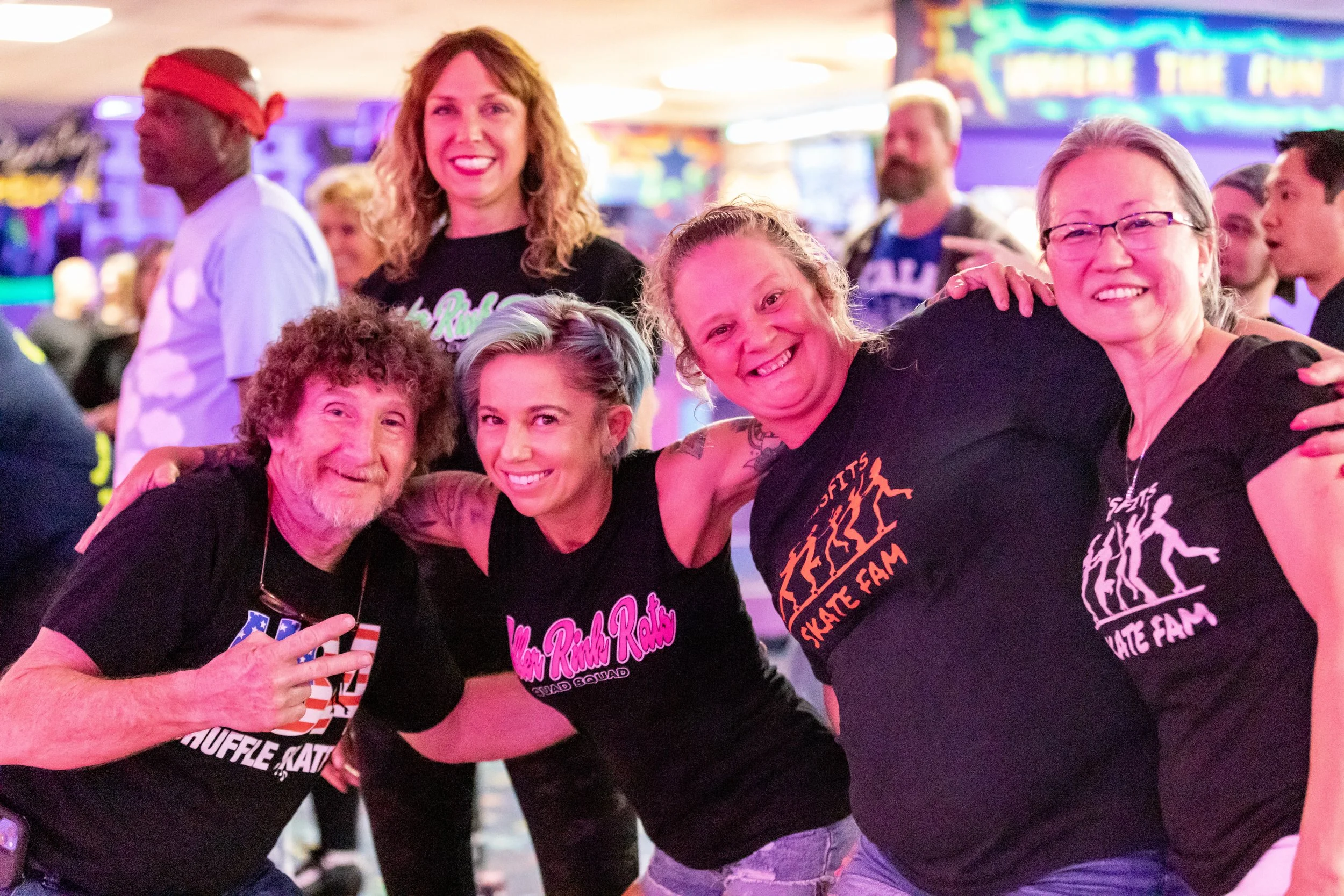 A group of five smiling people at a roller skating rink, some wearing roller skates and themed T-shirts, with colorful lights and a sign that says 'Hold the Fun' in the background.