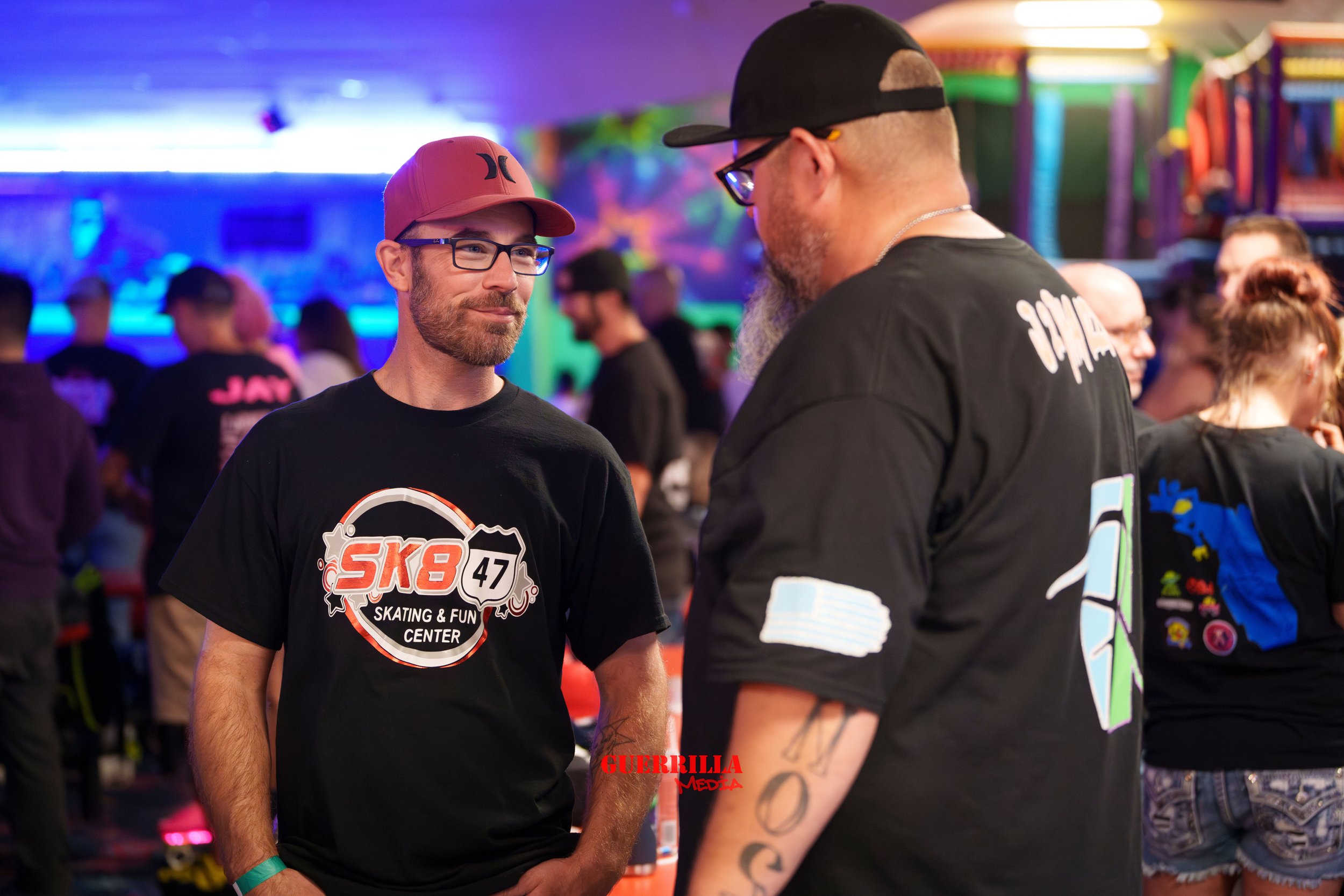 Two men wearing black T-shirts and glasses talking in a lively indoor roller skating rink with colorful lighting and other skaters in the background.