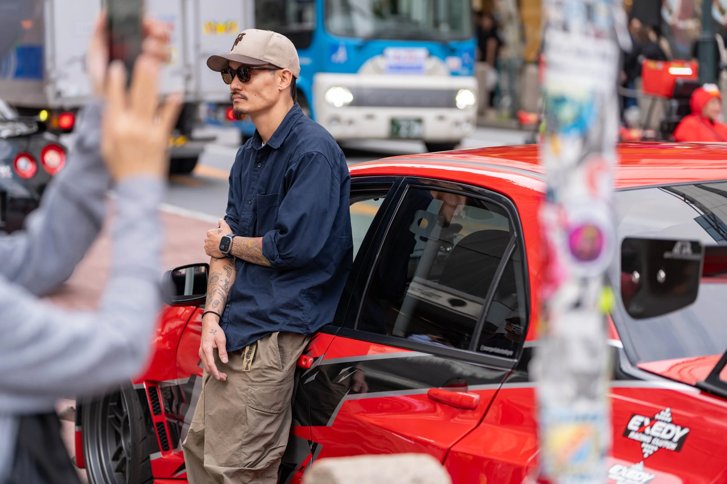 A man with tattoos on his left arm, wearing sunglasses, a tan cap, a navy blue shirt, and cargo pants, stands leaning against a red and black car on a city street. There are people and vehicles in the background.