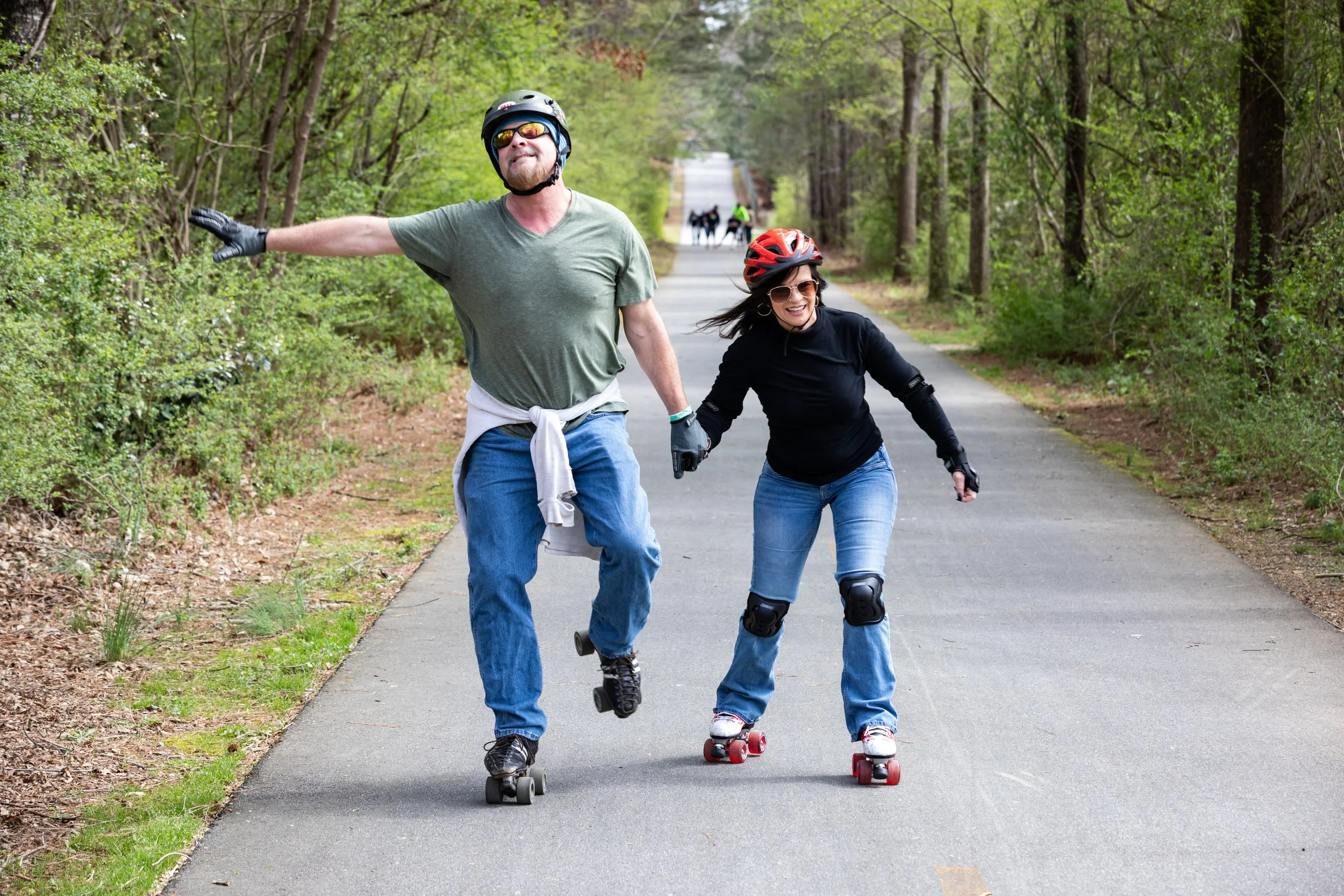 Two people roller skating on a paved trail in a forest, holding hands and smiling, with others skating in the background.