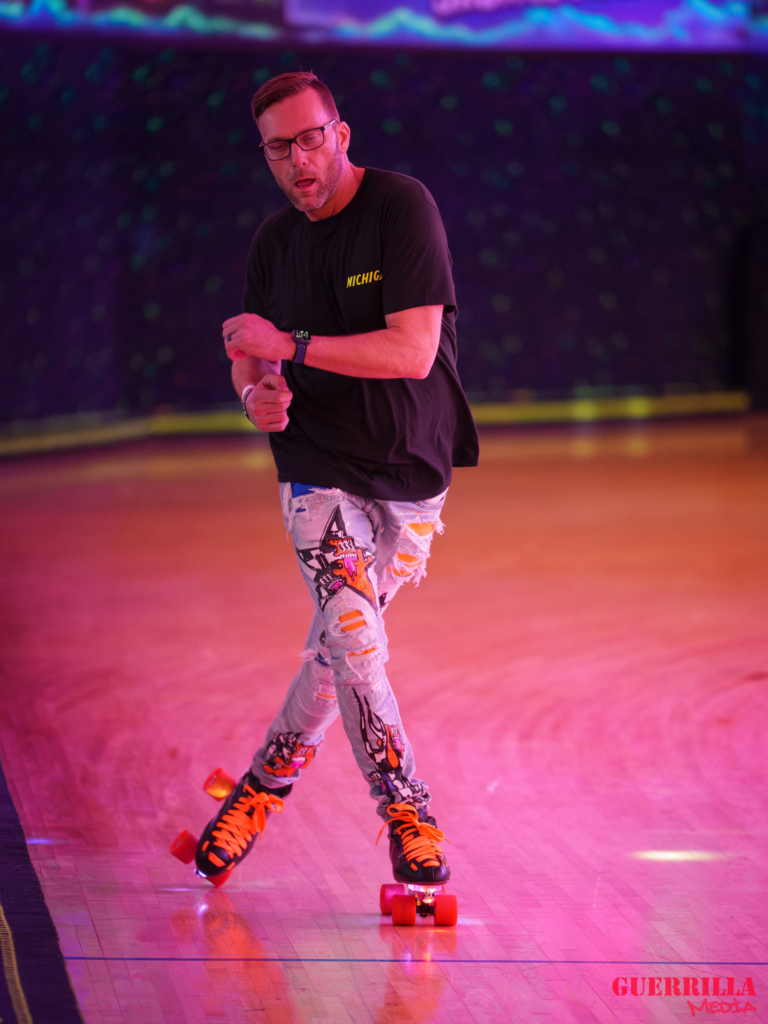 A man roller skating on a wooden rink with pinkish-purple lighting, wearing a black T-shirt and torn, colorful jeans.