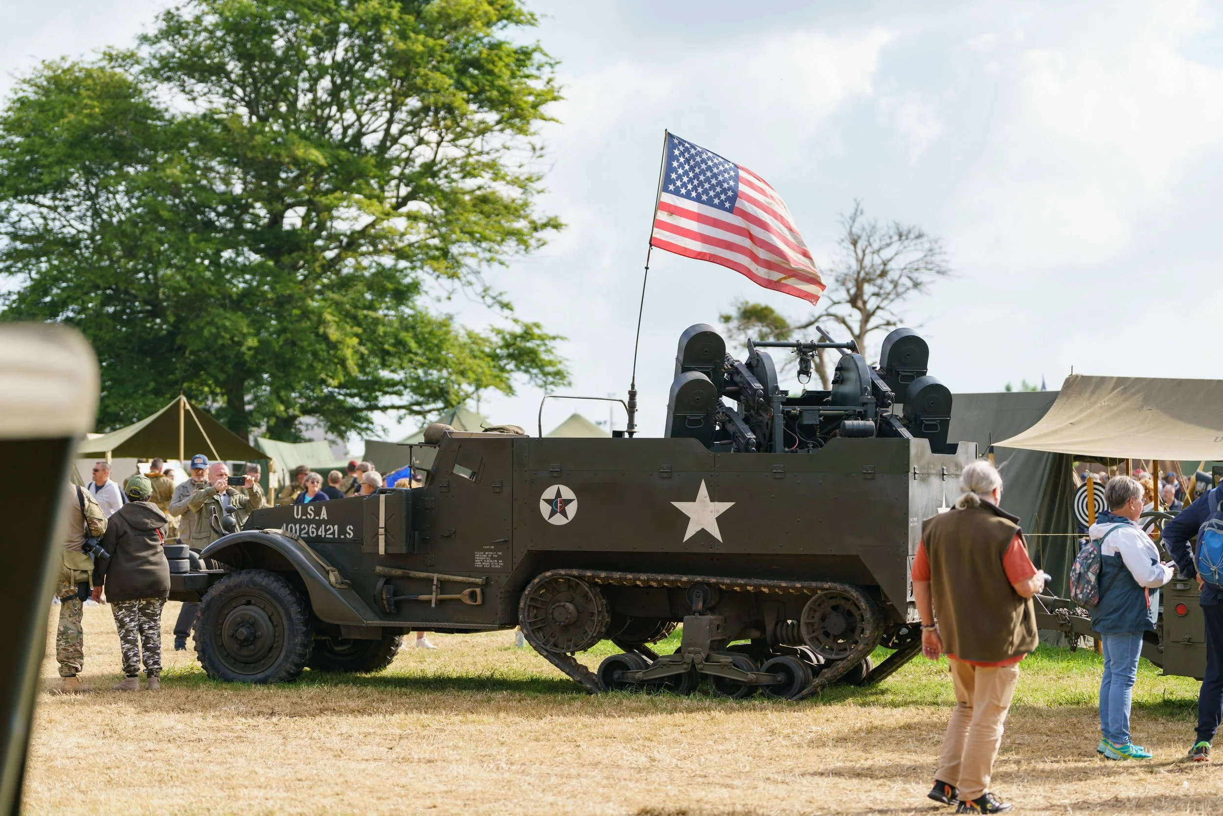 A vintage military tank with a U.S. flag mounted on top, surrounded by people at an outdoor event, with green trees and tents in the background.